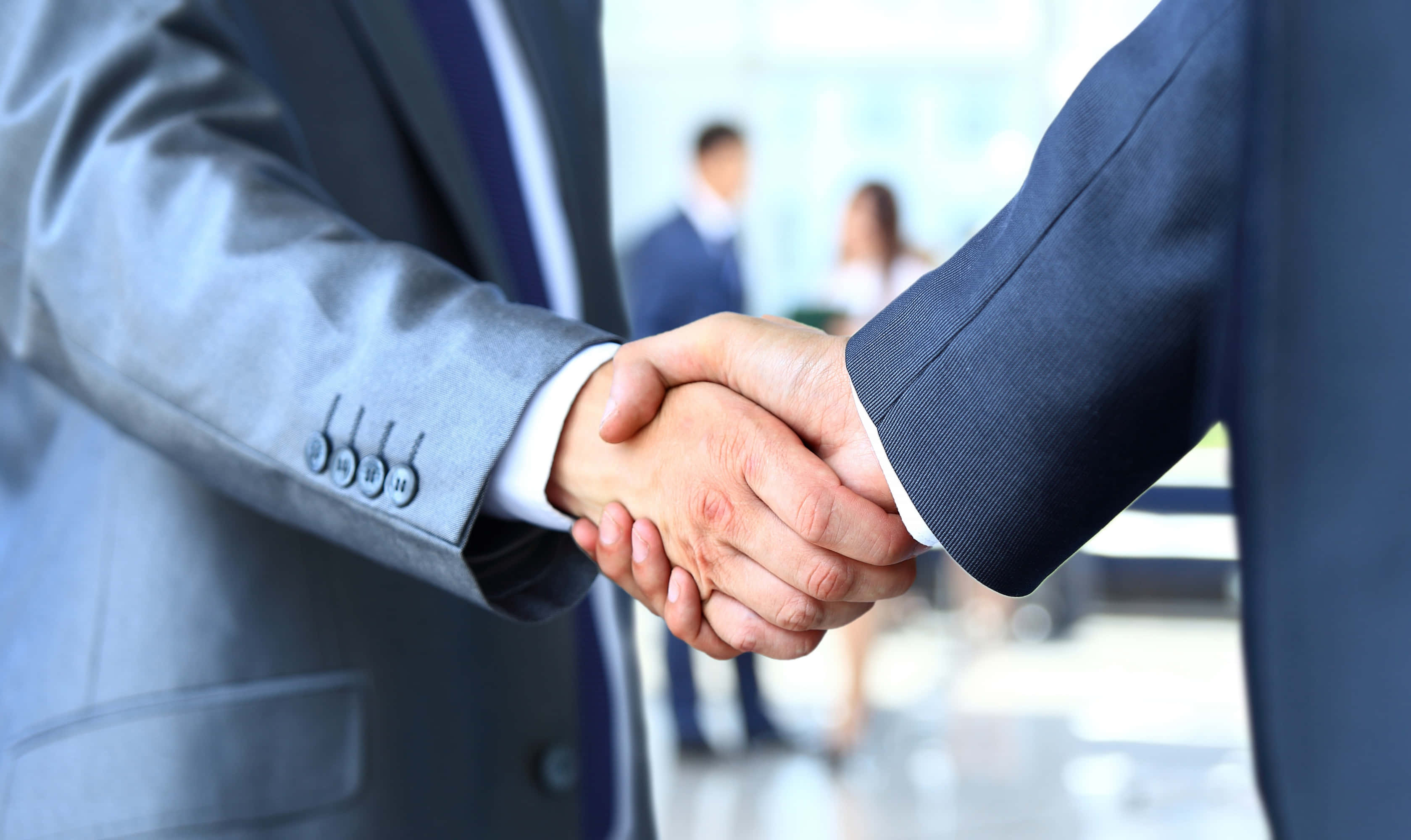 Businessman 桌布 壁紙 — 兩位身穿西裝的商務人士在明亮的現代辦公室中堅定握手，背景有模糊的同事。