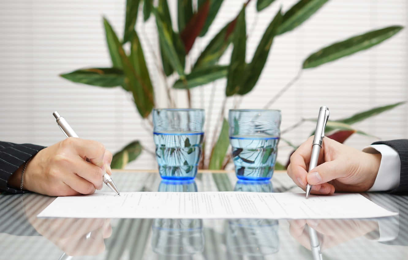 hands 桌布 壁紙 — 兩位身穿西裝的商務人士在玻璃桌上簽署合約，背景有玻璃水杯和一棵茂盛的植物。