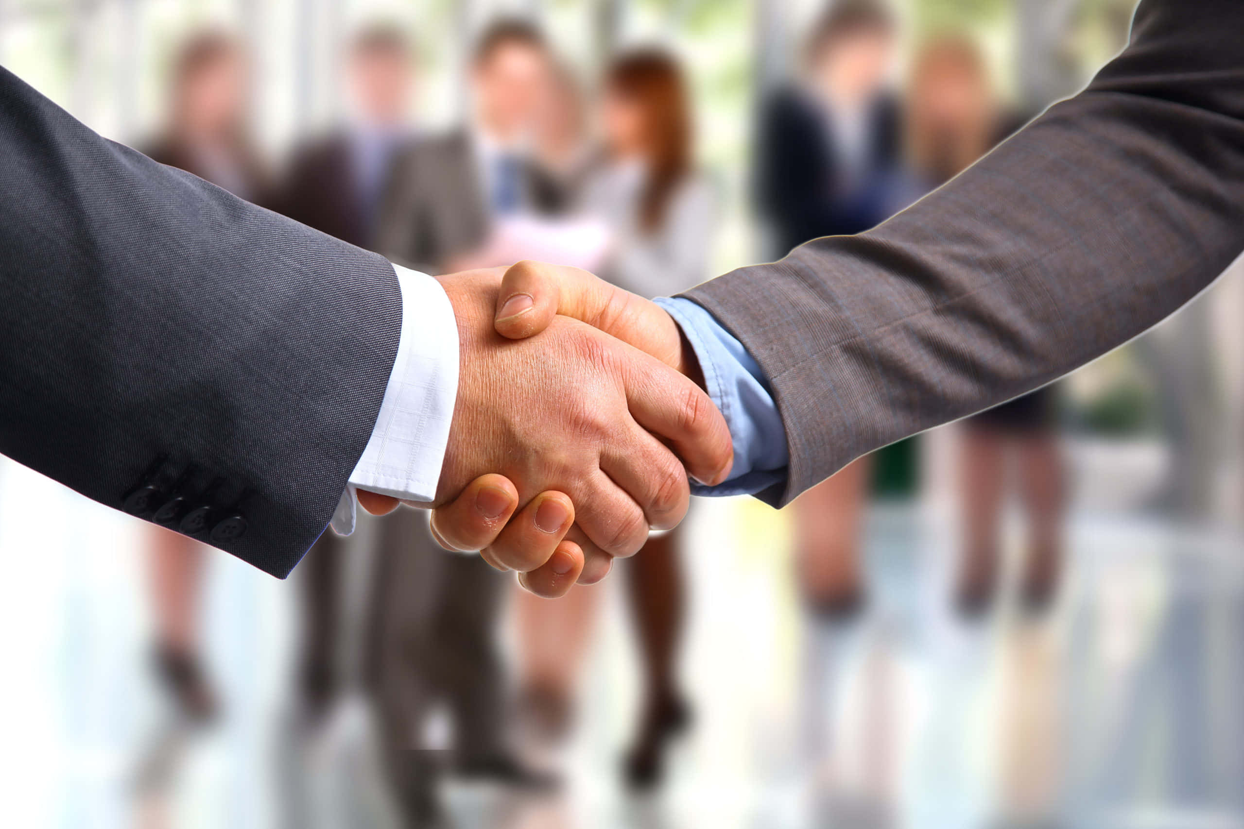 hands 桌布 壁紙 — 兩位身穿西裝的商務人士在現代辦公室中堅定握手，背景有模糊的同事。