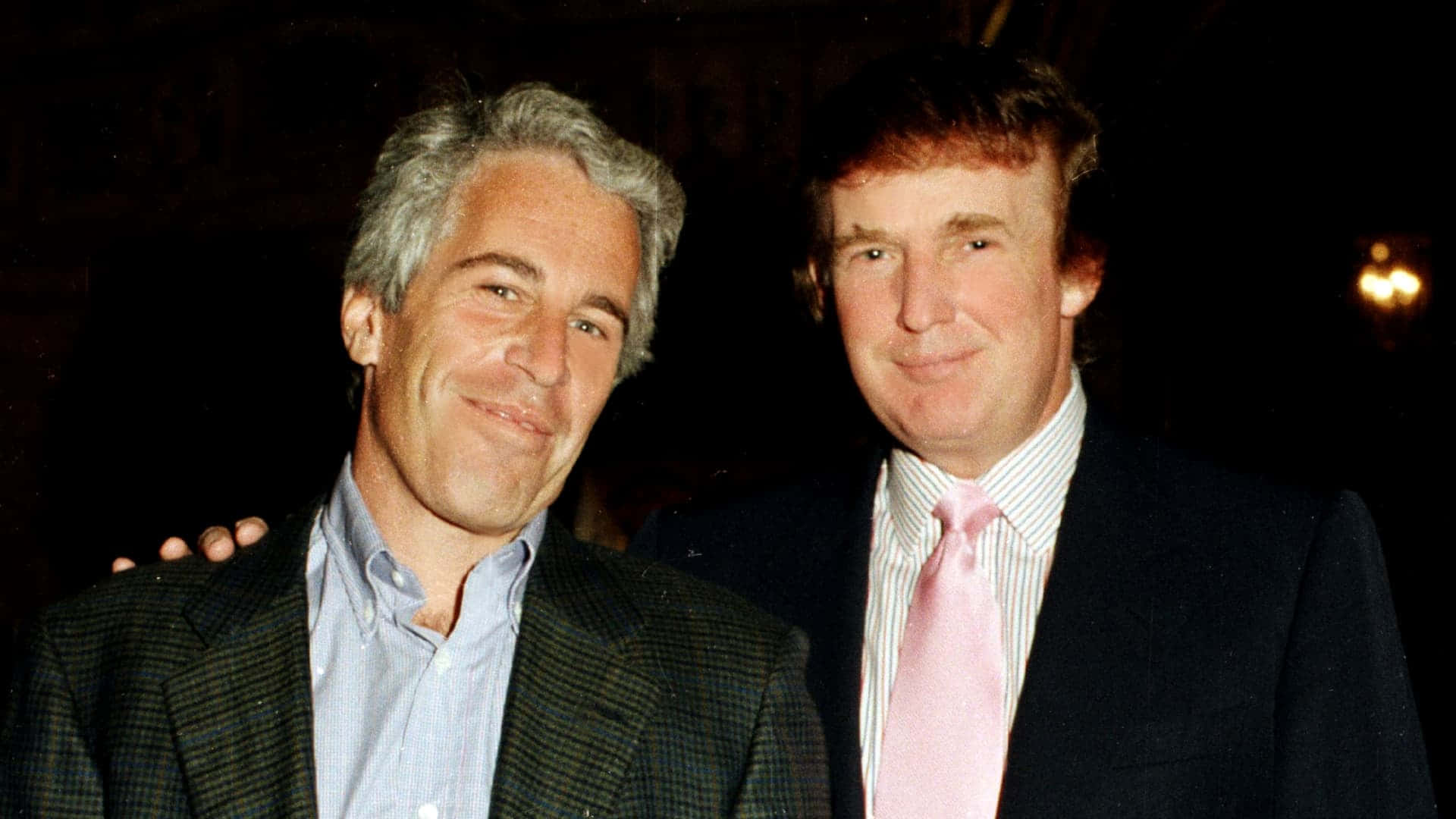 Bill Gates And Epstein 桌布 壁紙 — 兩位身穿西裝的男士在燈光昏暗、背景溫暖的晚間活動中緊密地擺姿勢。