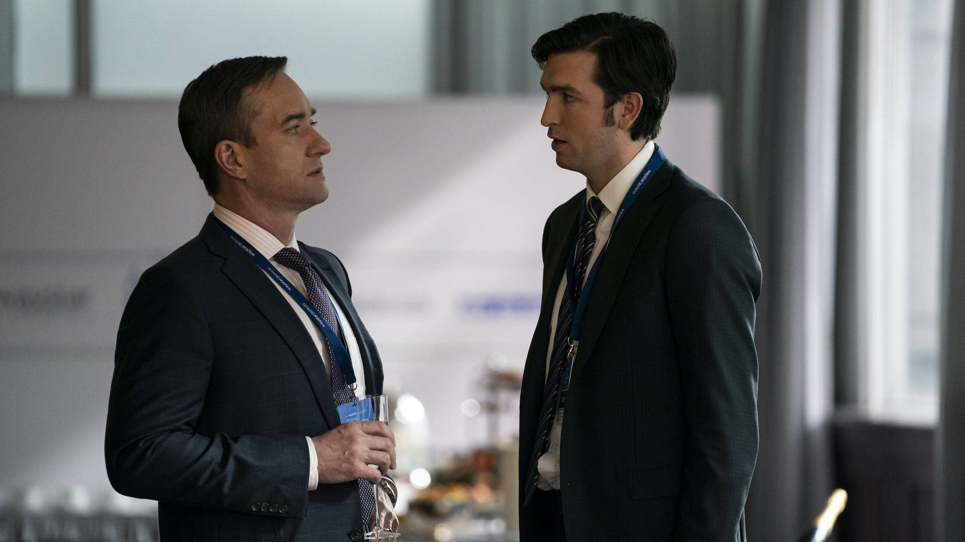 Succession 桌布 壁紙 — 兩位身穿西裝的男子在會議中專注交談，背景燈光模糊。