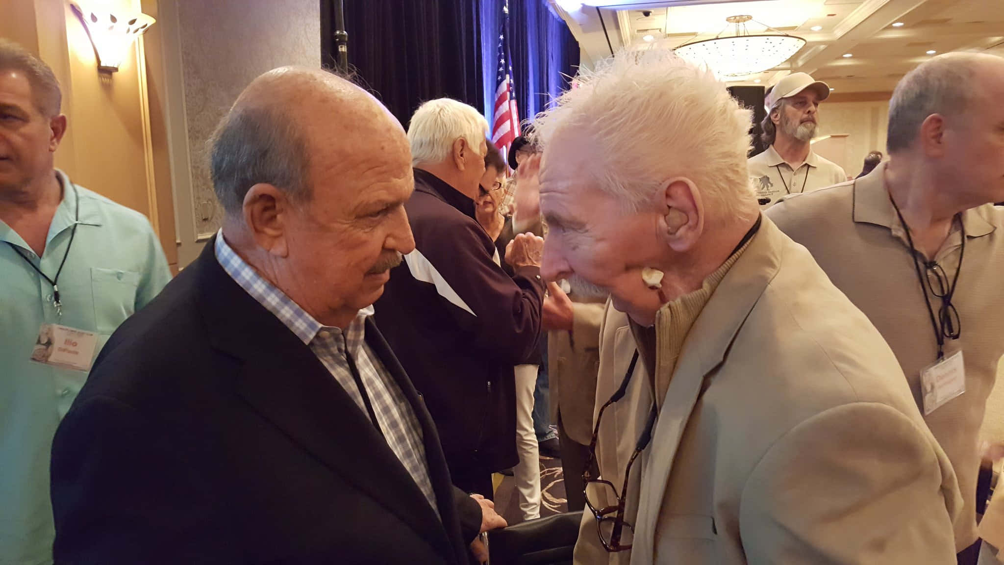 Bobby Heenan 桌布 壁紙 — 兩位身穿西裝的長者在一個擠滿人群的室內活動中，與美國國旗和吊燈一起分享溫暖的微笑時刻。
