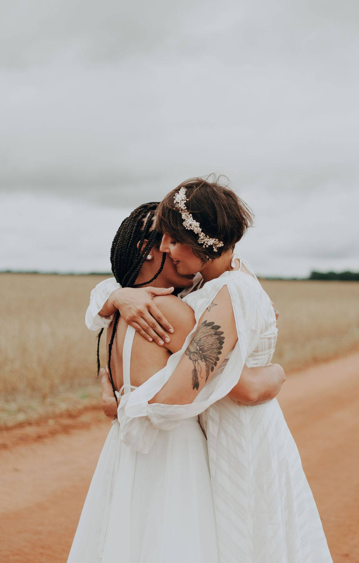 Subtle Lgbt 桌布 壁紙 — 兩位身穿飄逸白色連衣裙的女性在陰天下一條穿過金色田野的泥路上溫柔擁抱。