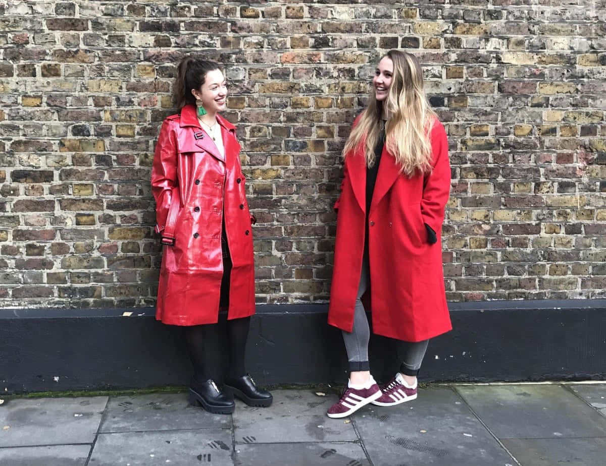 Red Coat 桌布 壁紙 — 兩位身穿鮮紅色外套的女性在城市的行人道上，對著一面風化的磚牆大笑。