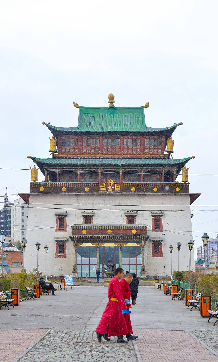 建築 桌布 壁紙 — 兩位身穿鮮紅色長袍的僧侶，在陰天之下走過一座宏偉的傳統蒙古寺廟，寺廟有綠色屋頂和金色裝飾。