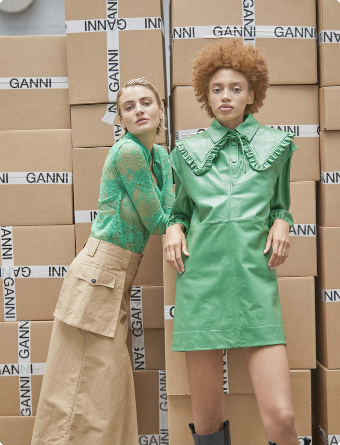 Green Fashion 桌布 壁紙 — 兩位身穿鮮綠色服裝的女性自信地擺姿勢，背景是堆疊的GANNI品牌紙箱牆。