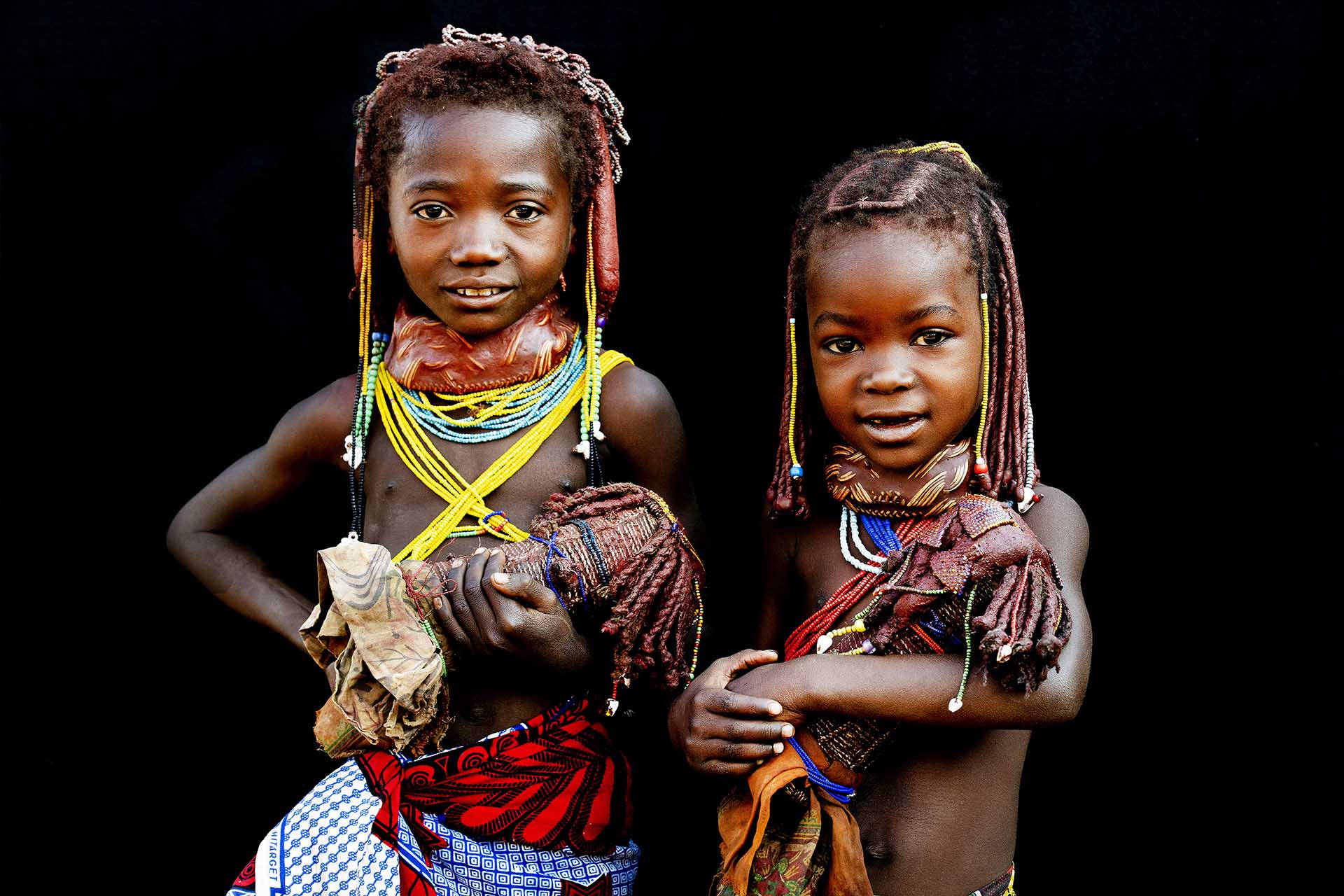 Angola 桌布 壁紙 — 兩位身穿鮮豔傳統服飾的年輕女孩在黑色背景前自信地擺姿勢。