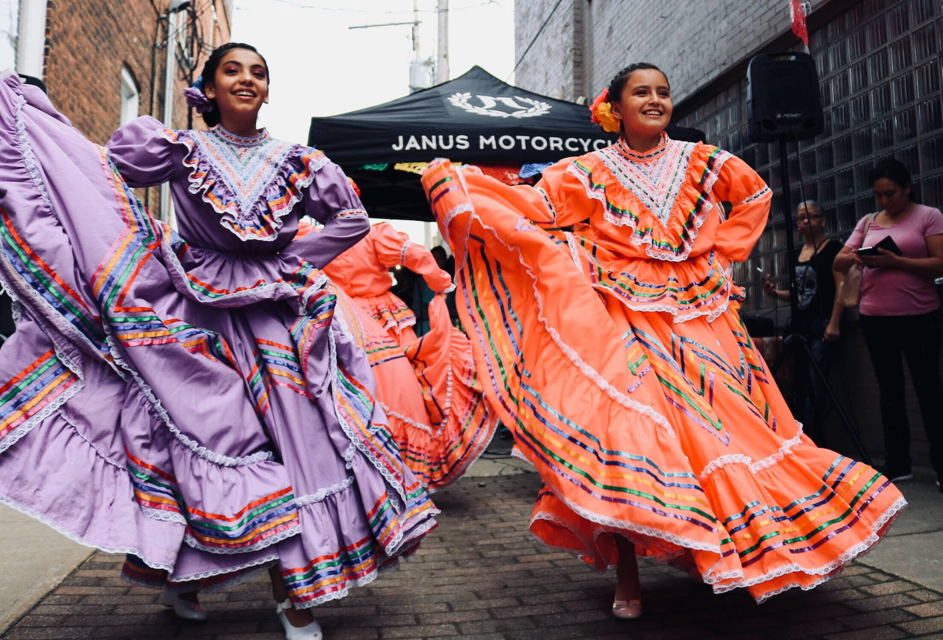 Mexican Woman 桌布 壁紙 — 兩位身穿鮮豔傳統服飾的快樂舞者，在熱鬧的文化慶典中，於磚砌街道上旋轉，裙擺飄逸。