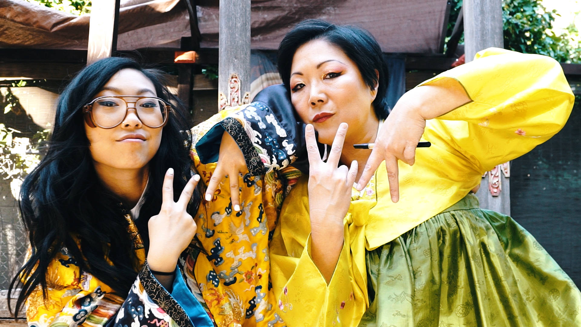 Awkwafina 桌布 壁紙 — 兩位身穿鮮豔傳統長袍的女性在明亮的日光下，俏皮地擺出和平手勢。
