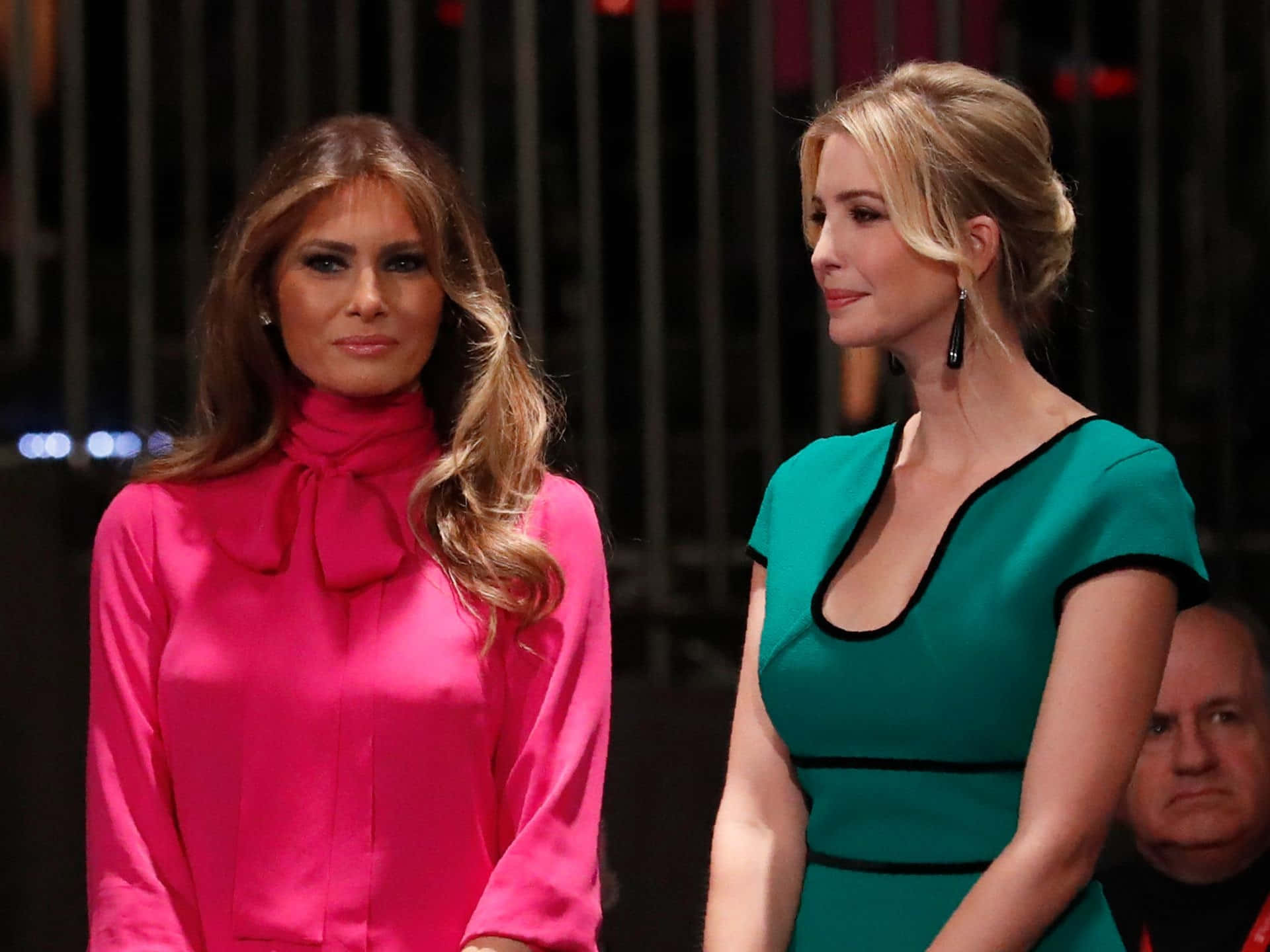 Melania Trump 桌布 壁紙 — 兩位身穿鮮豔粉紅和藍綠色連衣裙的女性在深色背景的正式活動中並肩站立。