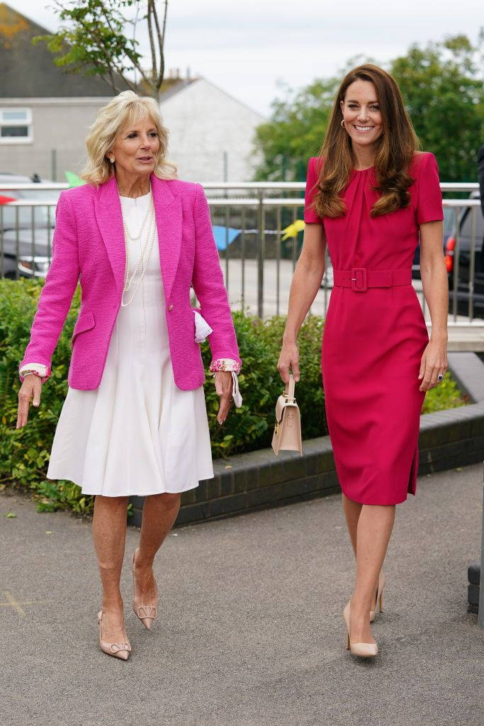 Kate Middleton 桌布 壁紙 — 兩位身穿鮮豔粉紅色服裝的女性並肩走在鋪砌的小路上，周圍環繞著綠植和建築物。