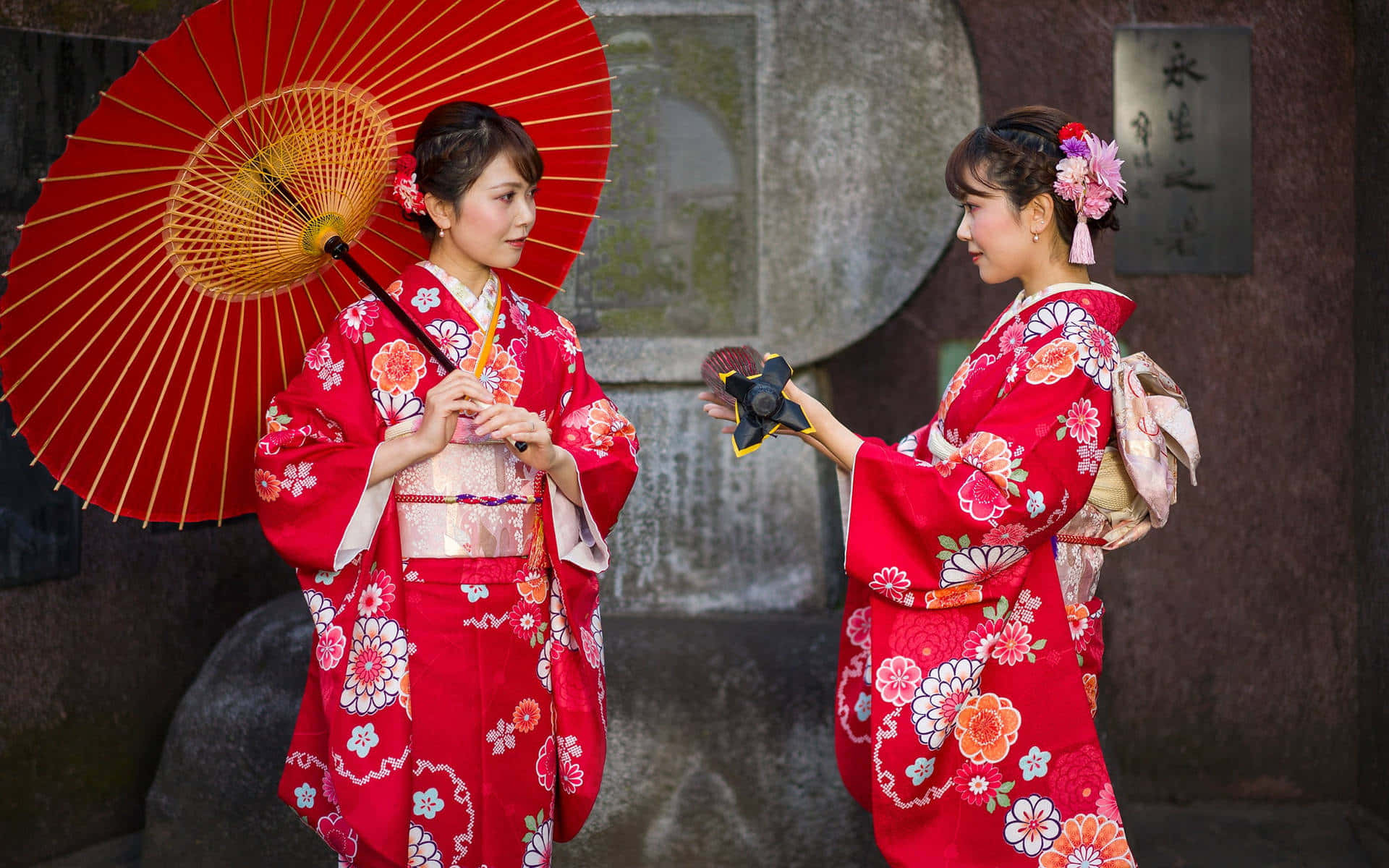 Japanese Women 桌布 壁紙 — 兩位身穿鮮豔紅色花卉和服的女士面對面站立，一位手持紅色陽傘，另一位手持裝飾扇，背景為石牆。