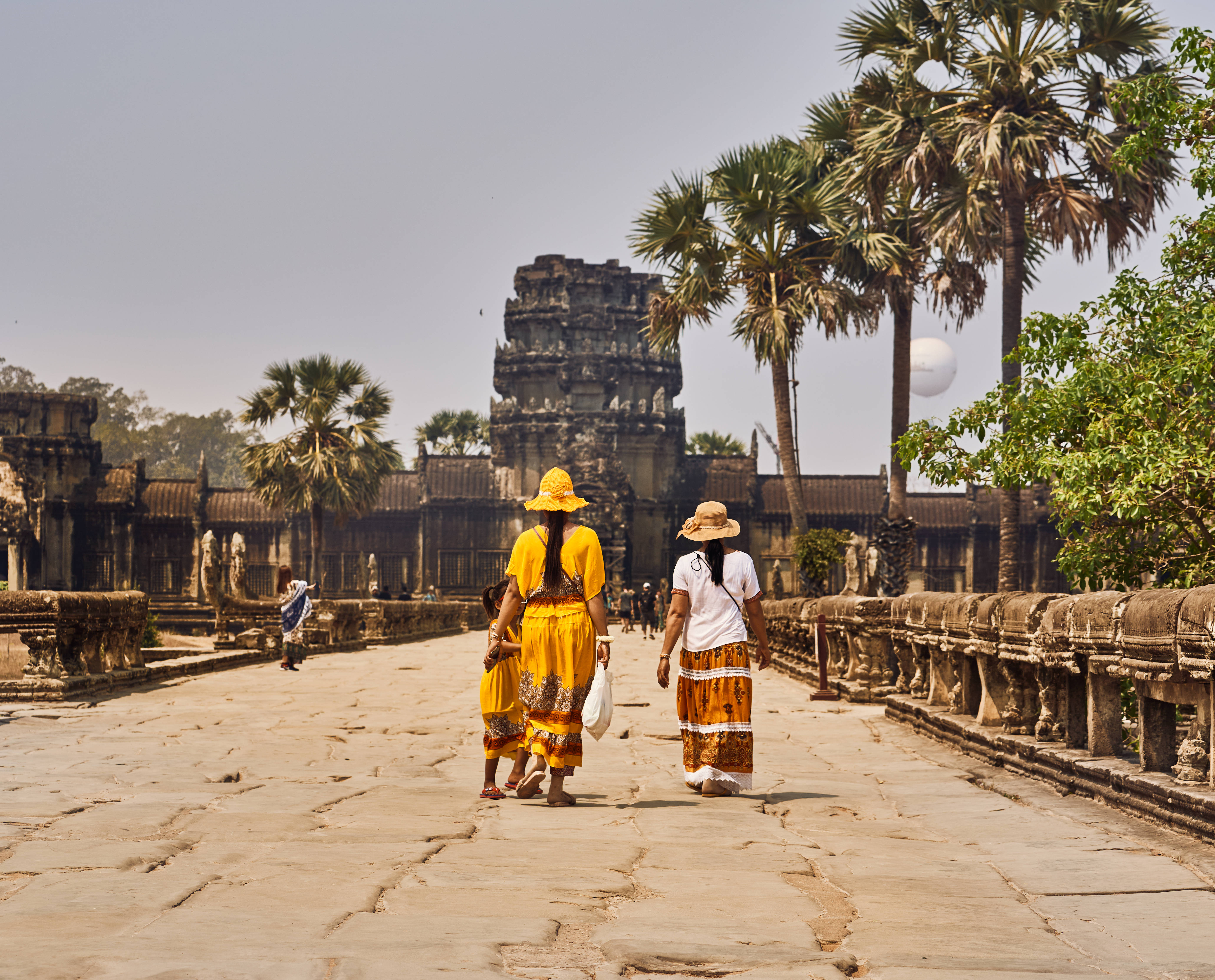 Cambodia 桌布 壁紙 — 兩位身穿鮮豔黃色和橙色服裝的女性，沿著石徑走向被高大棕櫚樹環繞的古老寺廟遺址，天空朦朧。