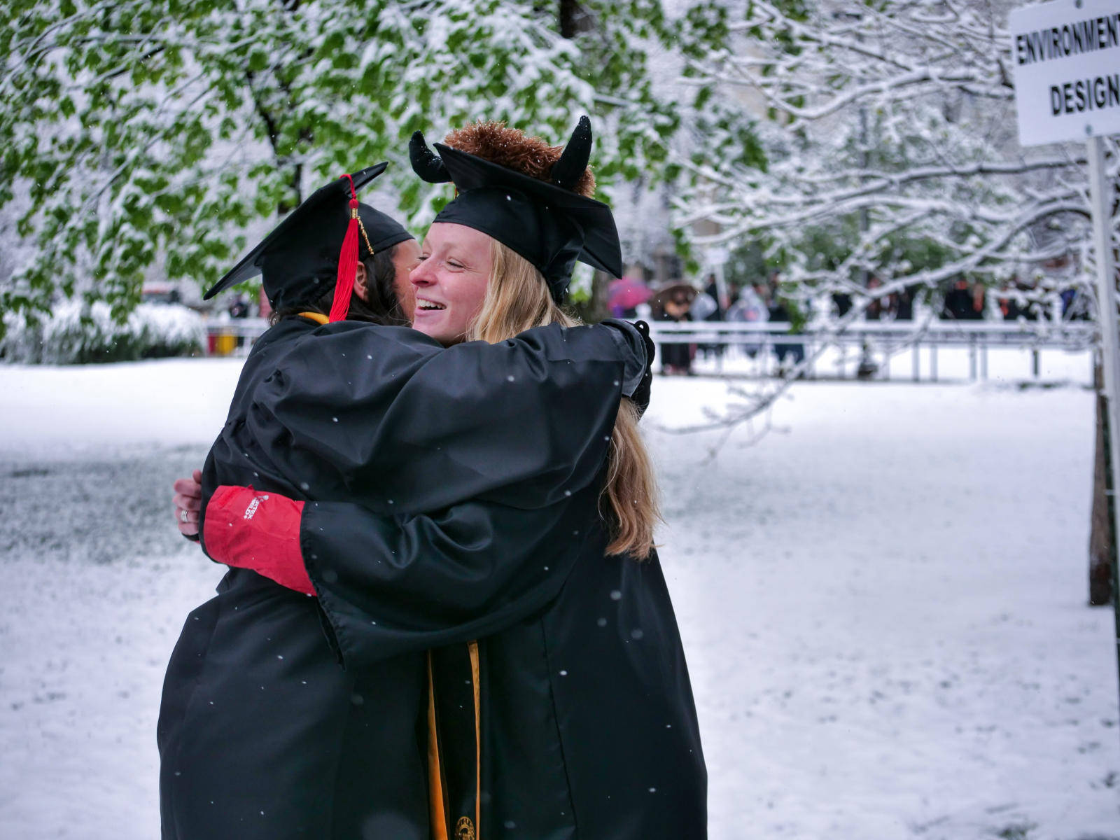 Graduation 桌布 壁紙 — 兩位身穿黑色學士帽和畢業袍的畢業生，在白雪覆蓋的戶外環境中熱情擁抱，背景有掛滿雪的樹木和一個寫著「ENVIRONMENT DESIGN」的標誌。