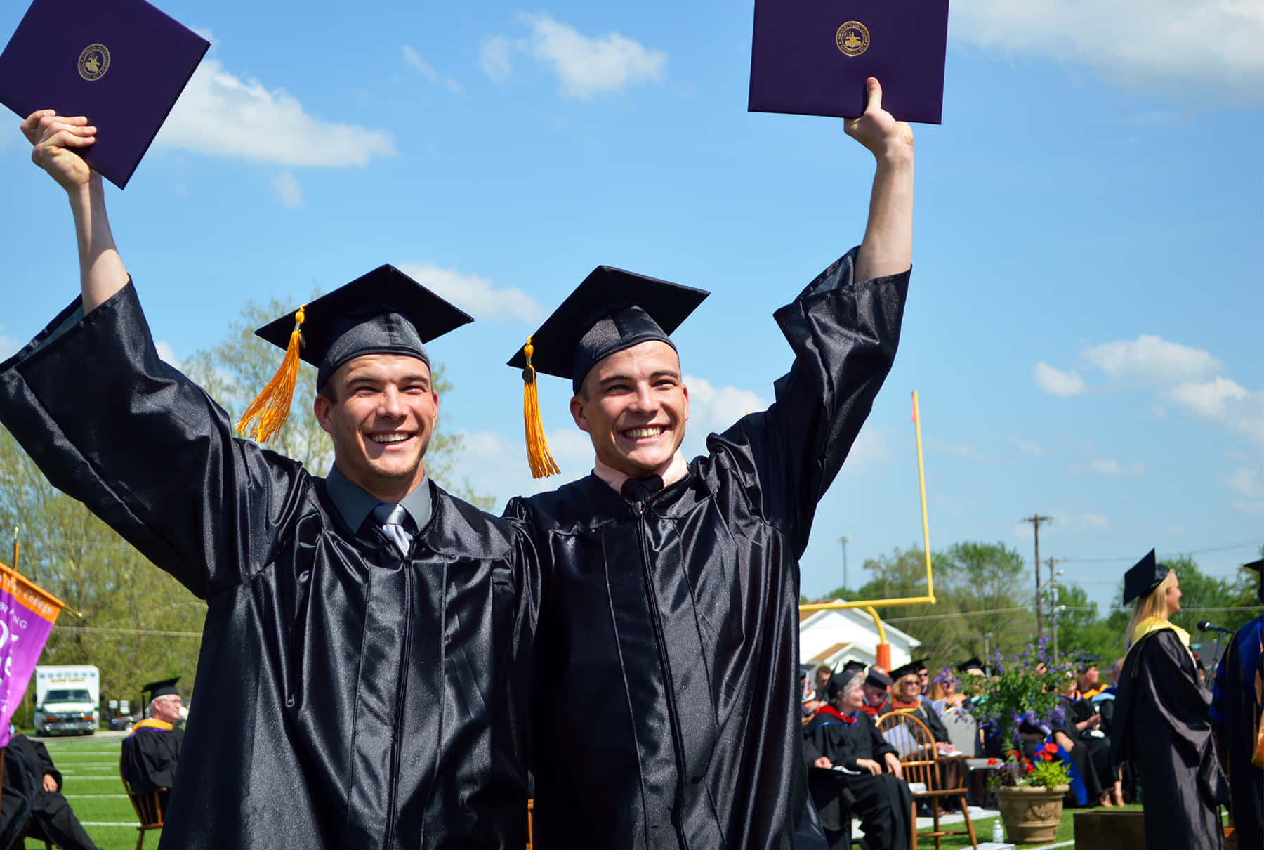 Graduation 桌布 壁紙 — 兩位身穿黑色學士袍和四方帽的快樂畢業生，在晴朗藍天下於戶外典禮中高舉文憑。