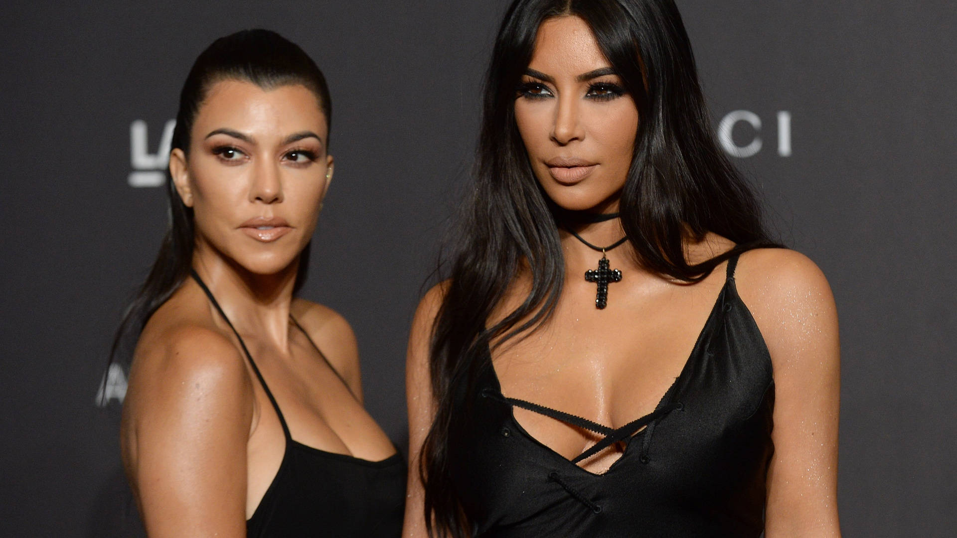 Kim Kardashian 桌布 壁紙 — 兩位身穿黑色晚裝的女性在一個華麗的活動中，自信地在深色品牌背景前擺姿勢。
