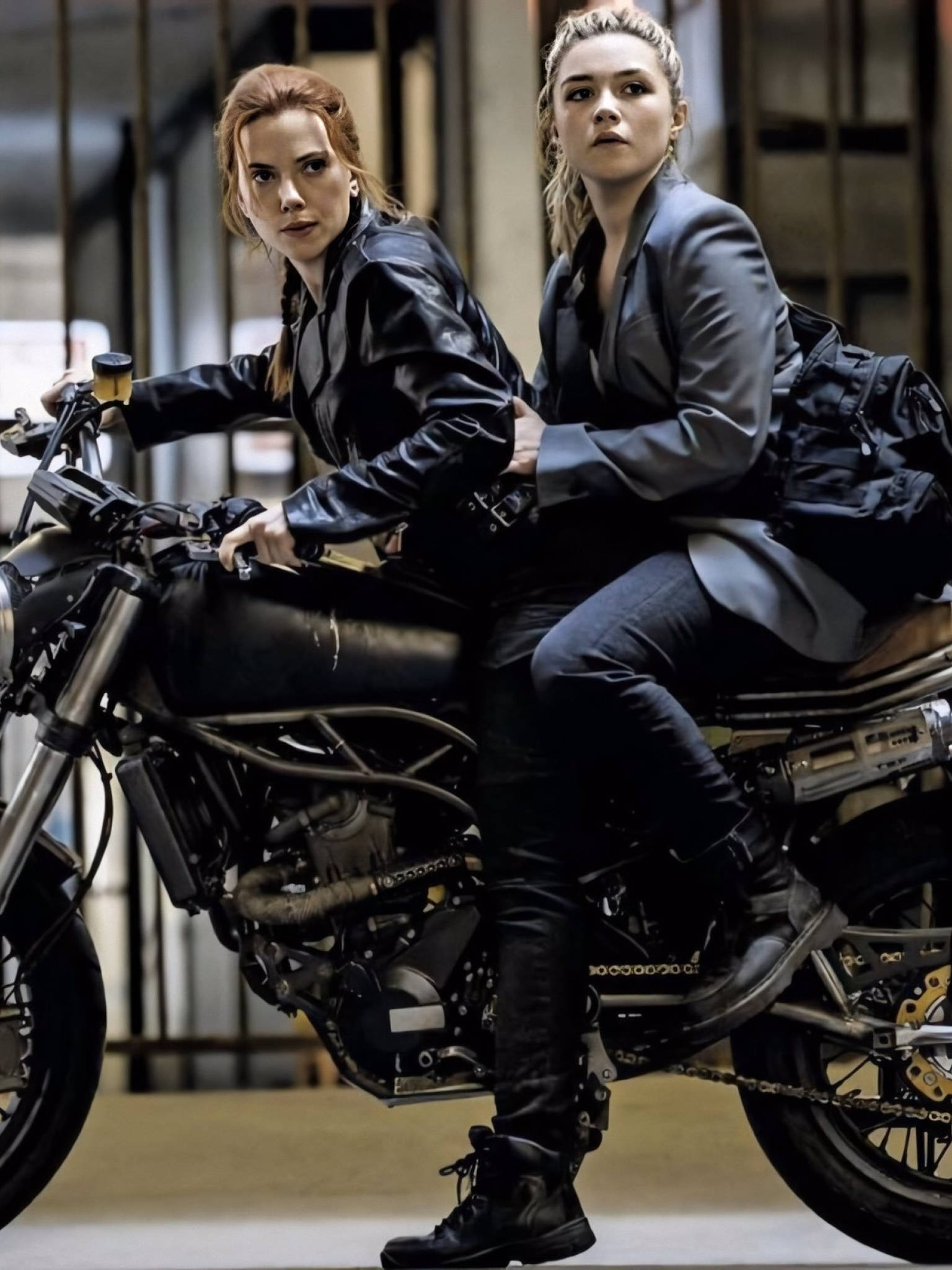 Scarlett Johansson Black Widow 桌布 壁紙 — 兩位身穿黑色皮革和西裝的女性，騎著堅固的摩托車穿過一個有鋼樑和昏暗照明的工業倉庫。