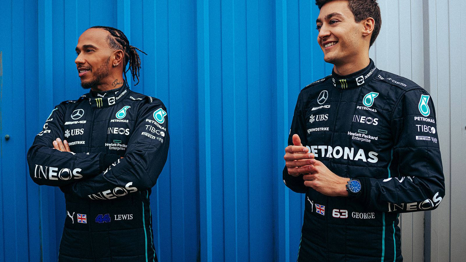 Mercedes 桌布 壁紙 — 兩位身穿黑色賽車服的F1車手，在充滿活力的藍色波紋牆前微笑站立。