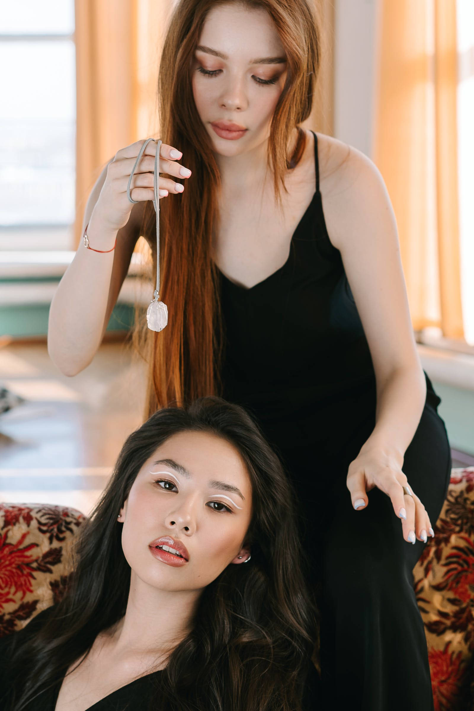 Hypnosis 桌布 壁紙 — 兩位身穿黑色連衣裙的女性，一位坐在花卉沙發上，另一位站著手持水晶吊墜，室內場景。