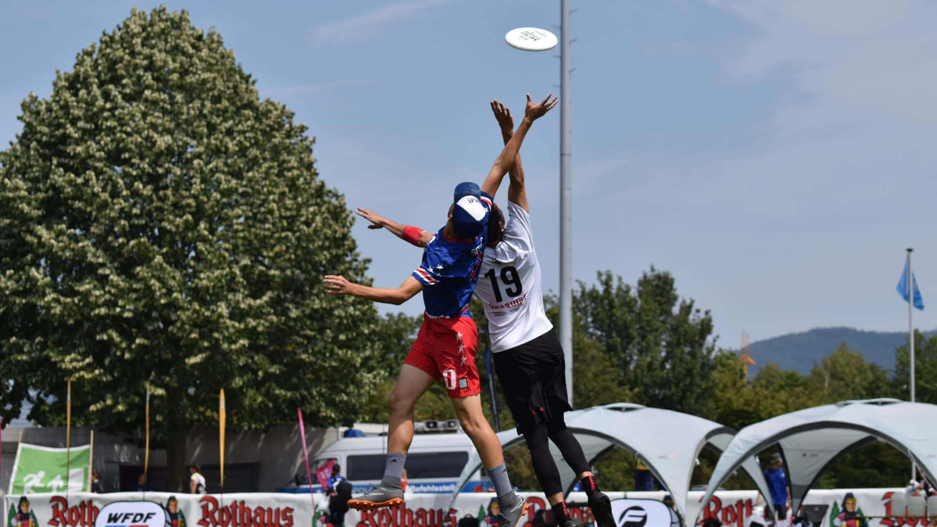 HD Frisbee 桌布 壁紙 — 兩位運動員在晴朗藍天下，背景有樹木和活動帳篷，跳起並伸手迎向飛行的飛盤。