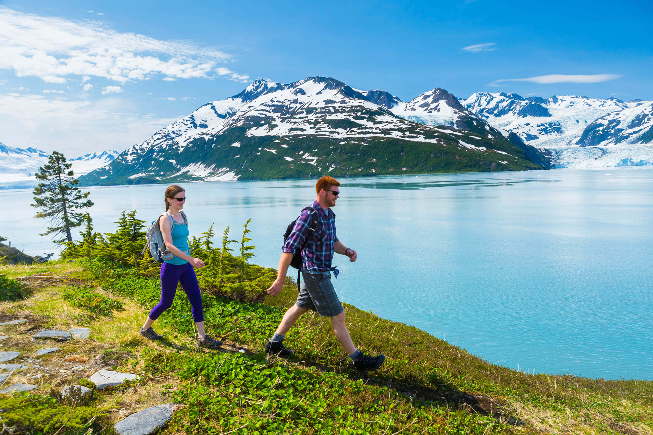 Eco-tourism 桌布 壁紙 — 兩位遠足人士在明亮的藍天下，沿著一條青草小徑漫步，旁邊是一個碧綠色的湖泊，遠處有積雪的山峰和冰川。