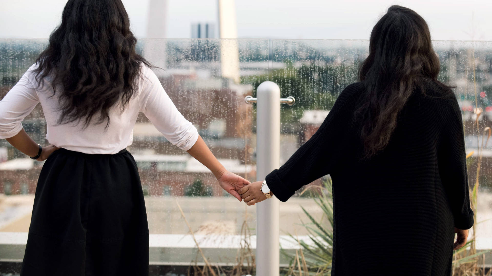 Bestie 桌布 壁紙 — 兩位長黑髮女性在雨中屋頂從後牽手，俯瞰城市景觀。