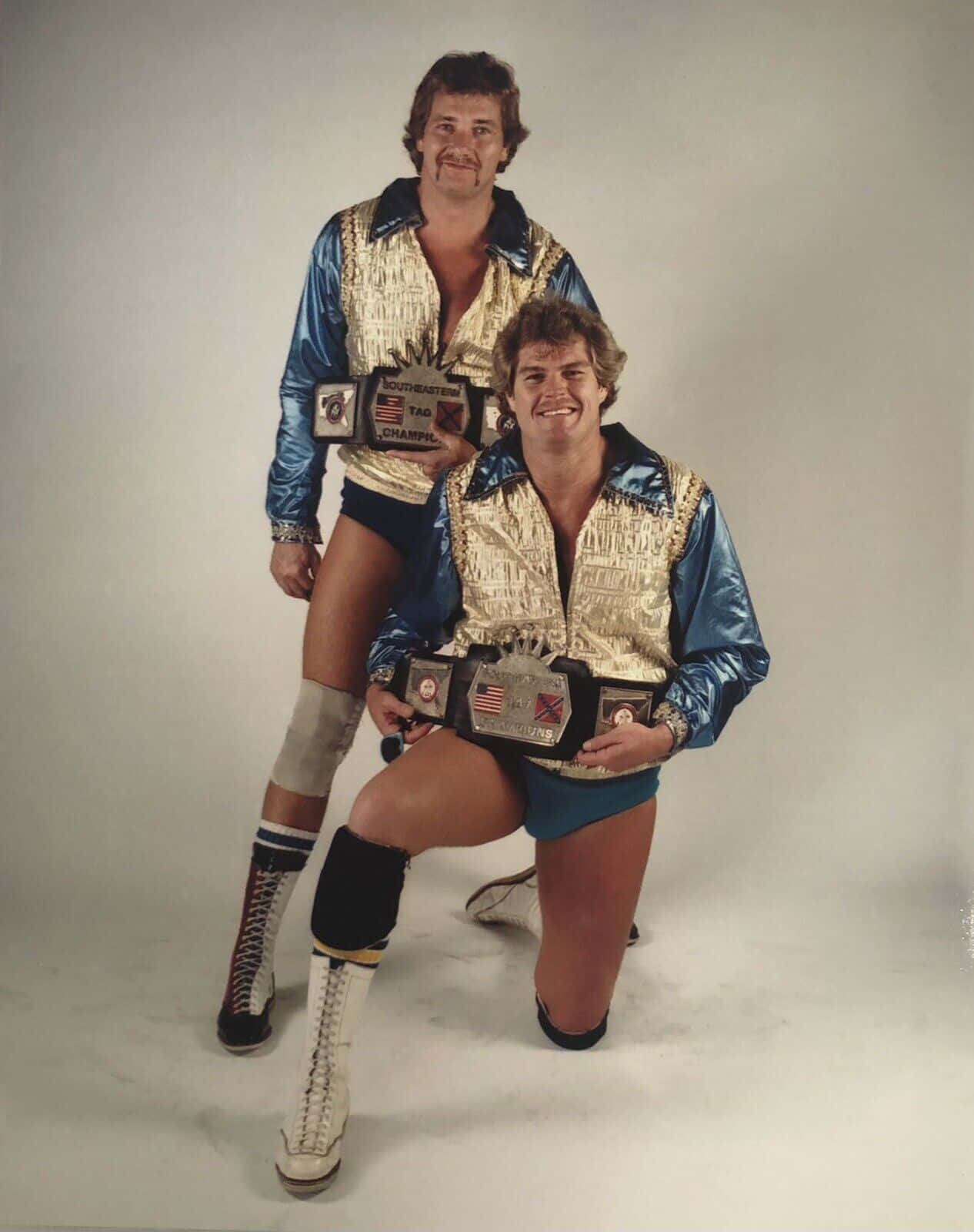 WWE 桌布 壁紙 — 兩位面帶微笑的1980年代摔角手，身穿閃亮藍色和金色外套，在樸素的工作室背景前自豪地展示冠軍腰帶。