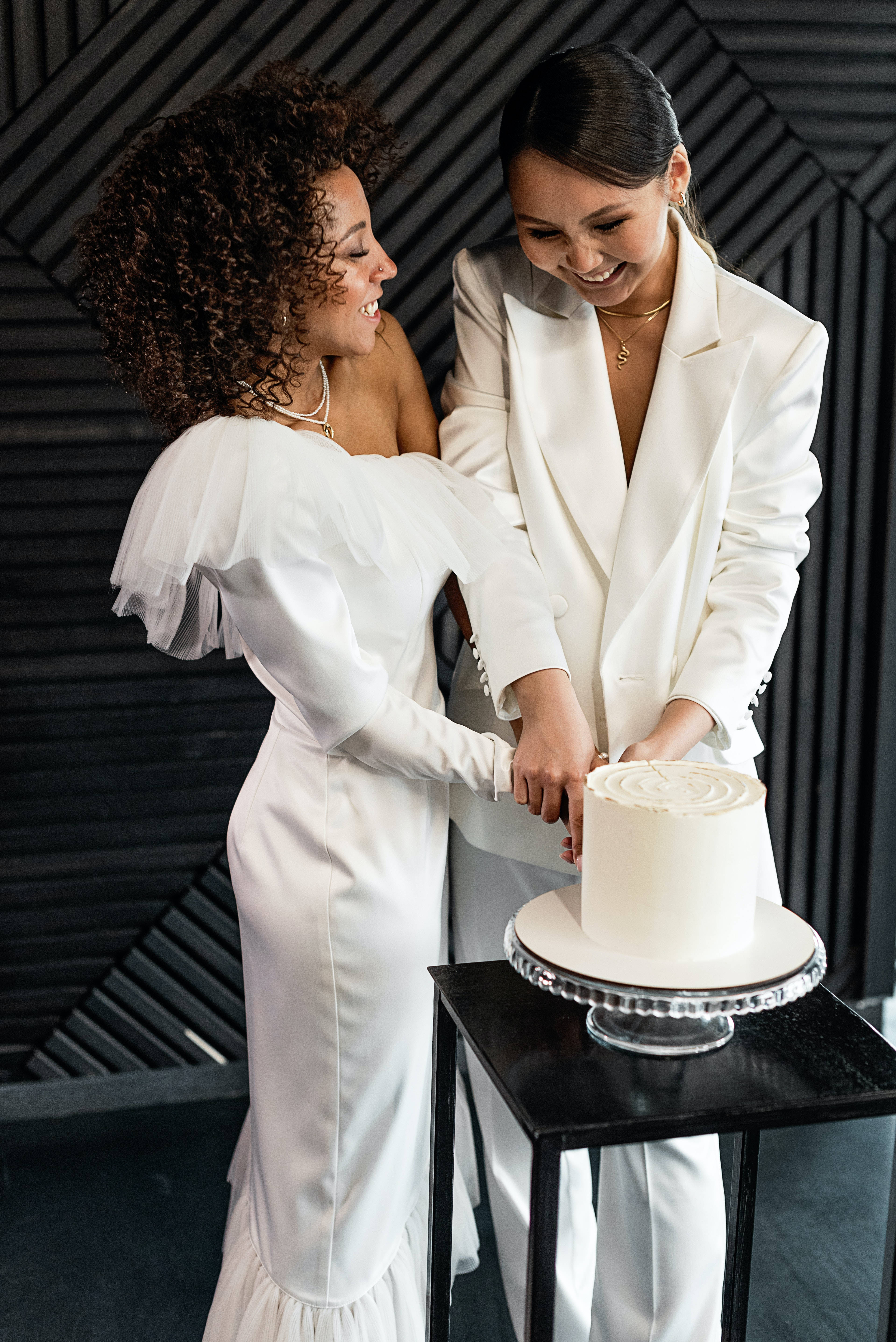 Wedding 桌布 壁紙 — 兩位面帶微笑的新娘身穿白色婚紗，在深色幾何牆前一起愉快地切白色蛋糕。