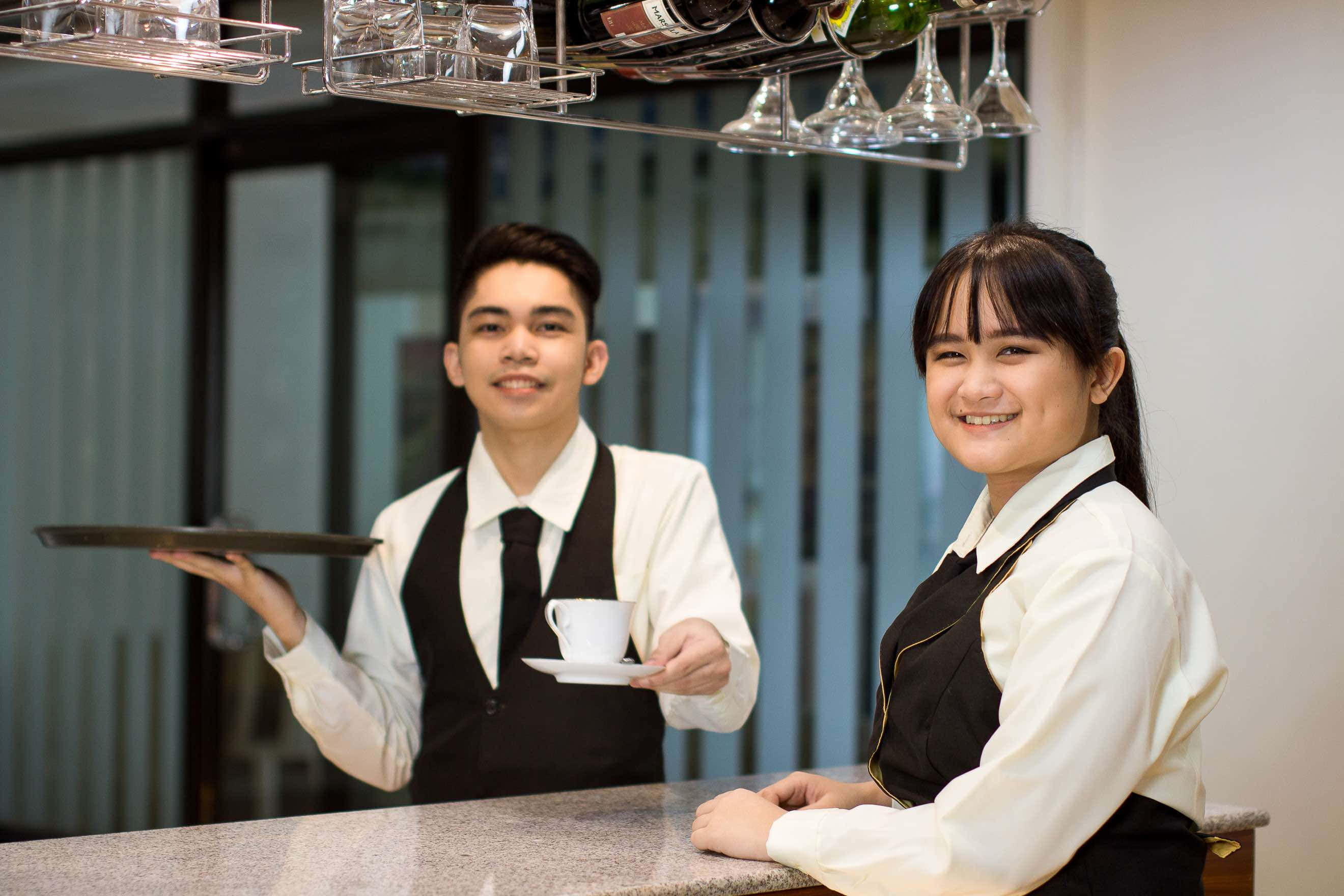 Hospitality 桌布 壁紙 — 兩位面帶微笑的服務生穿著整潔制服，在現代飯店大廳酒吧提供茶和葡萄酒。