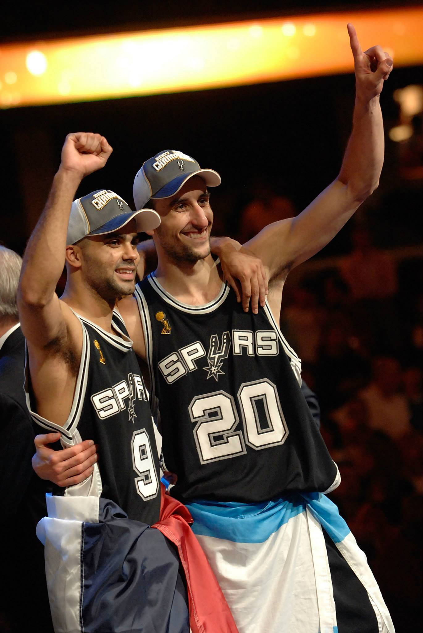 Manu 桌布 壁紙 — 兩位面帶微笑的聖安東尼奧馬刺籃球運動員穿著冠軍球衣，在場館燈光下高舉拳頭並戴上冠軍帽慶祝。