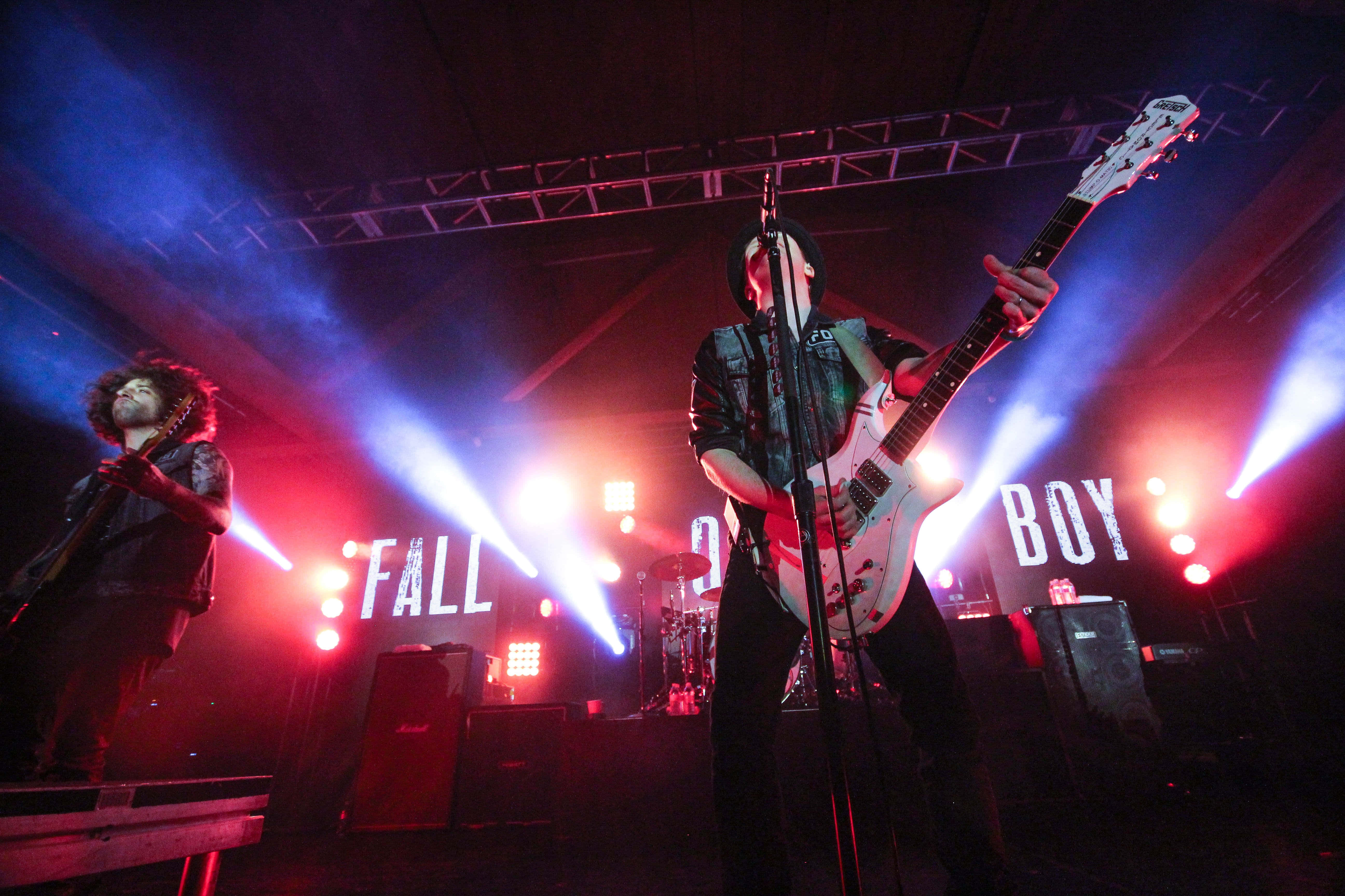 fall out boy 桌布 壁紙 — 兩位音樂家在昏暗的舞台上表演，舞台被紅色和藍色聚光燈籠罩，身後有「FALL OUT BOY」字樣亮起。
