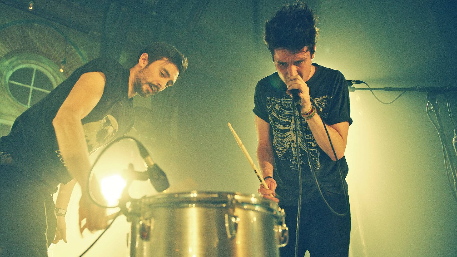 Bastille 桌布 壁紙 — 兩位音樂家在煙霧瀰漫、燈光昏暗的場地中，在戲劇性的舞台燈光下，與鼓手和主唱一起激烈表演。
