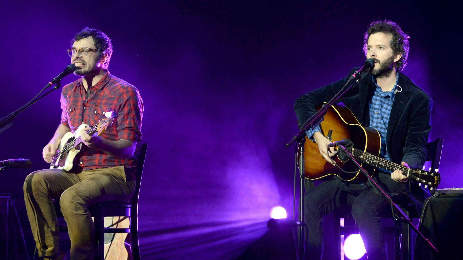 Flight Of The Conchords 桌布 壁紙 — 兩位音樂家在紫色燈光下於舞台上表演，一位彈奏低音吉他，另一位彈奏木吉他。