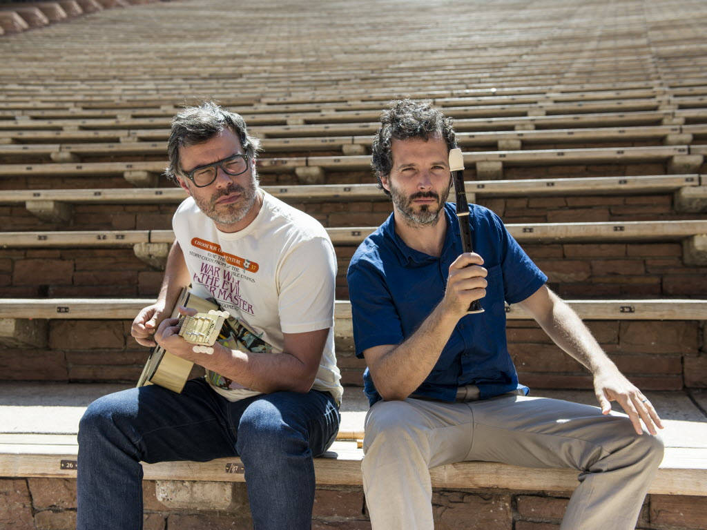 Flight Of The Conchords 桌布 壁紙 — 兩位音樂家坐在古老圓形劇場的石階上，一人手持吉他，另一人手持直笛，沐浴在明亮的陽光中。