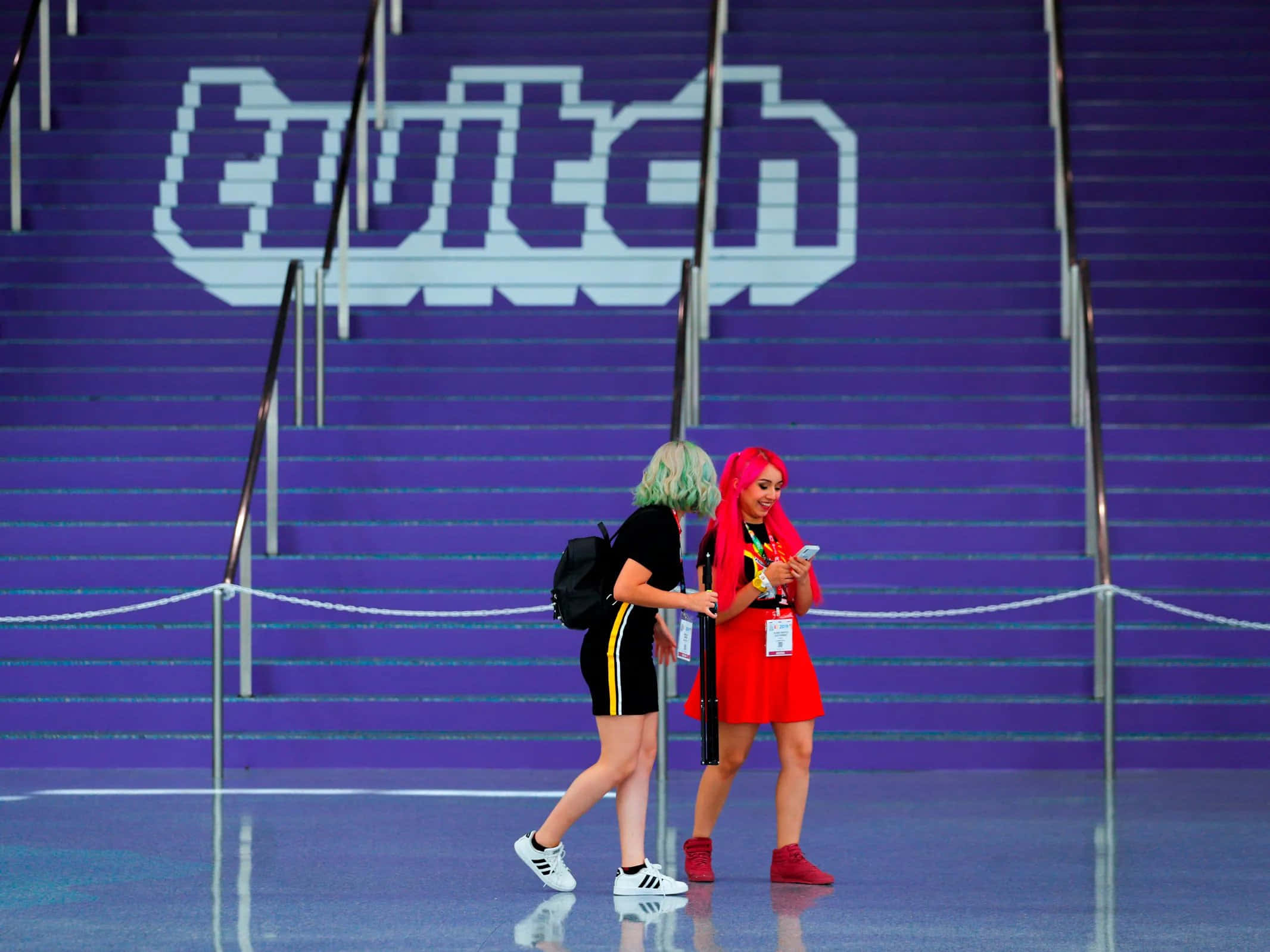 Twitch 桌布 壁紙 — 兩位頭髮鮮豔的會議參加者走過紫色樓梯上一個巨大的像素化Twitch標誌。
