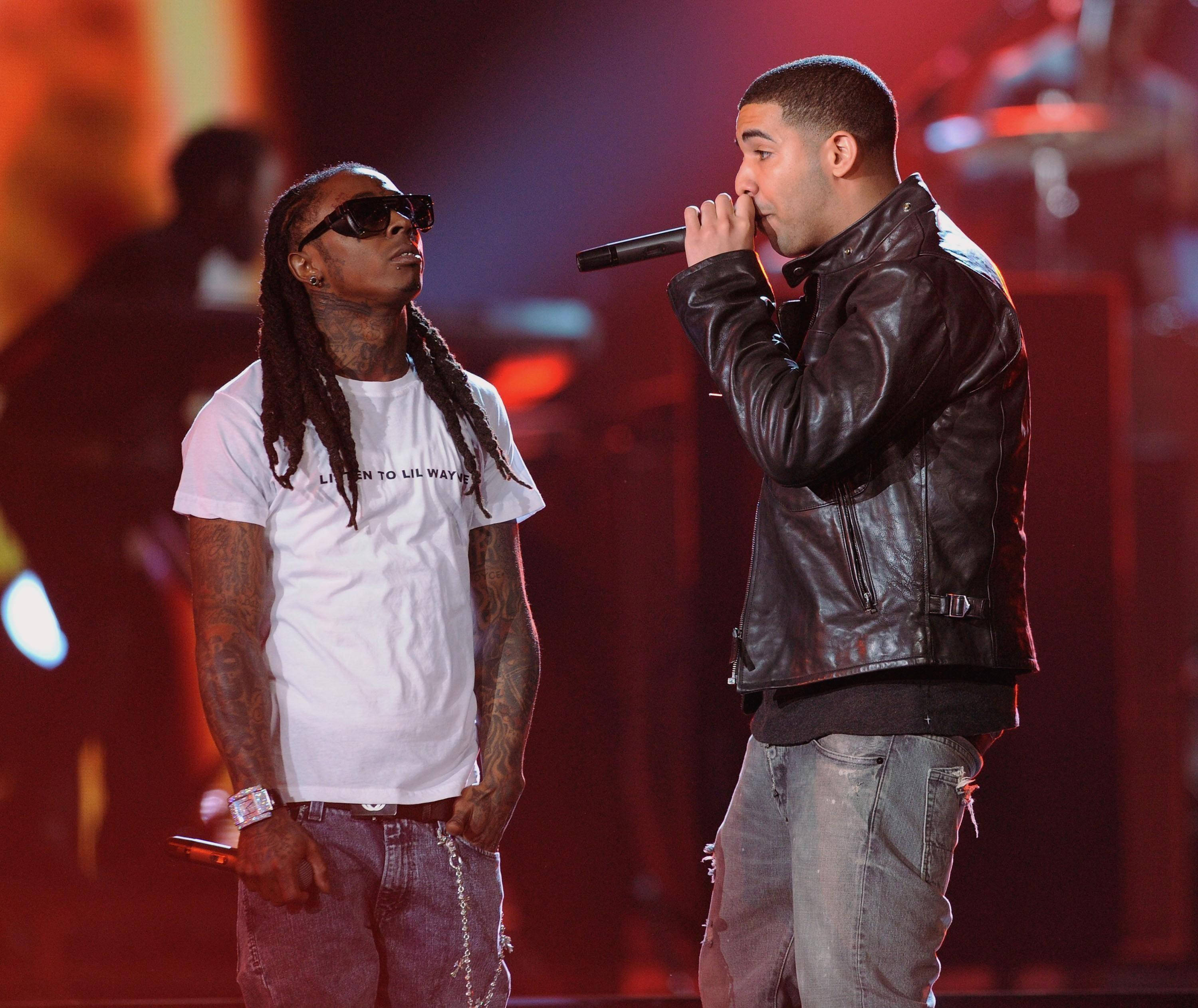 Lil Wayne 桌布 壁紙 — 兩位饒舌歌手在舞台上，一位身穿白色T恤和太陽眼鏡，另一位身穿皮夾克，在紅燈下對著麥克風唱歌。