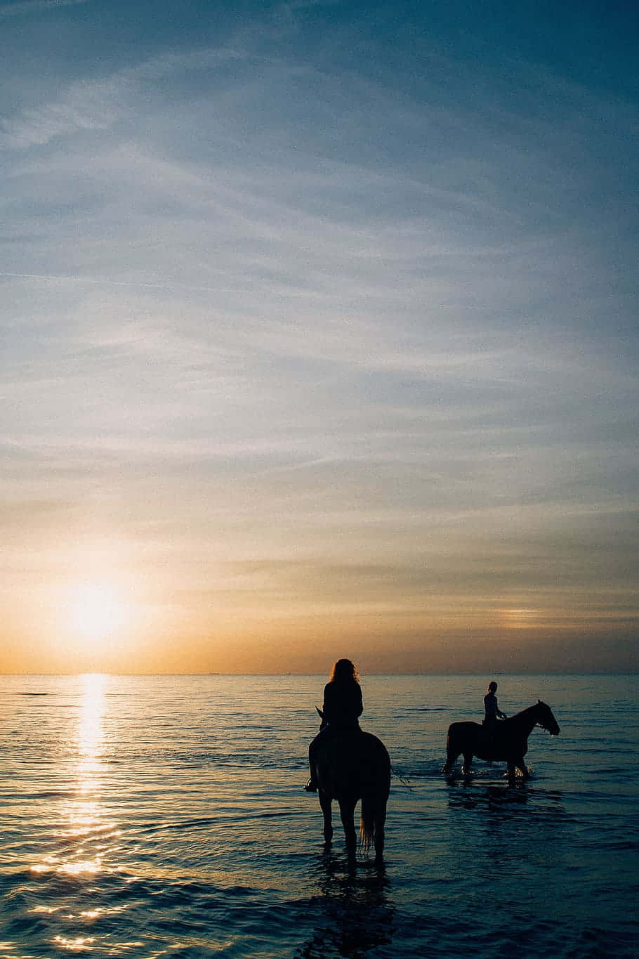 海灘 桌布 壁紙 — 兩位騎手騎著馬在淺水中涉水而行，夕陽西下，金色光芒灑在平靜的海面上。