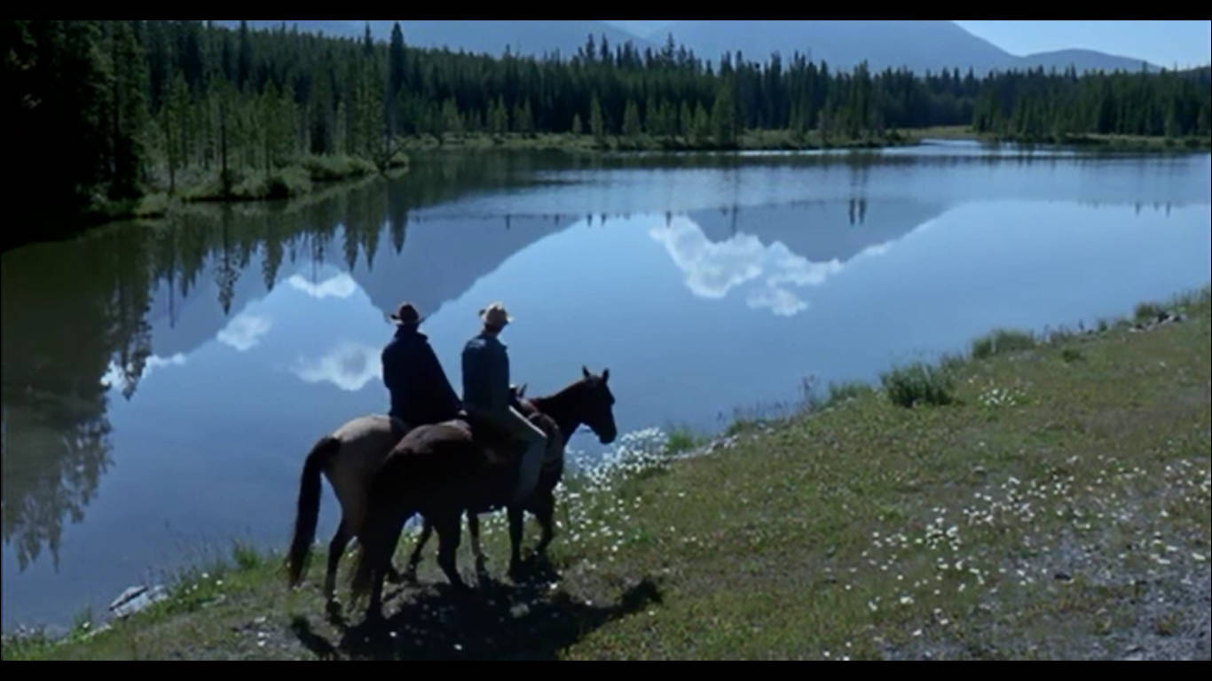 brokeback mountain 桌布 壁紙 — 兩位騎馬者在平靜的湖邊稍作停留，湖面倒映著雲朵和森林覆蓋的山脈，陽光普照。