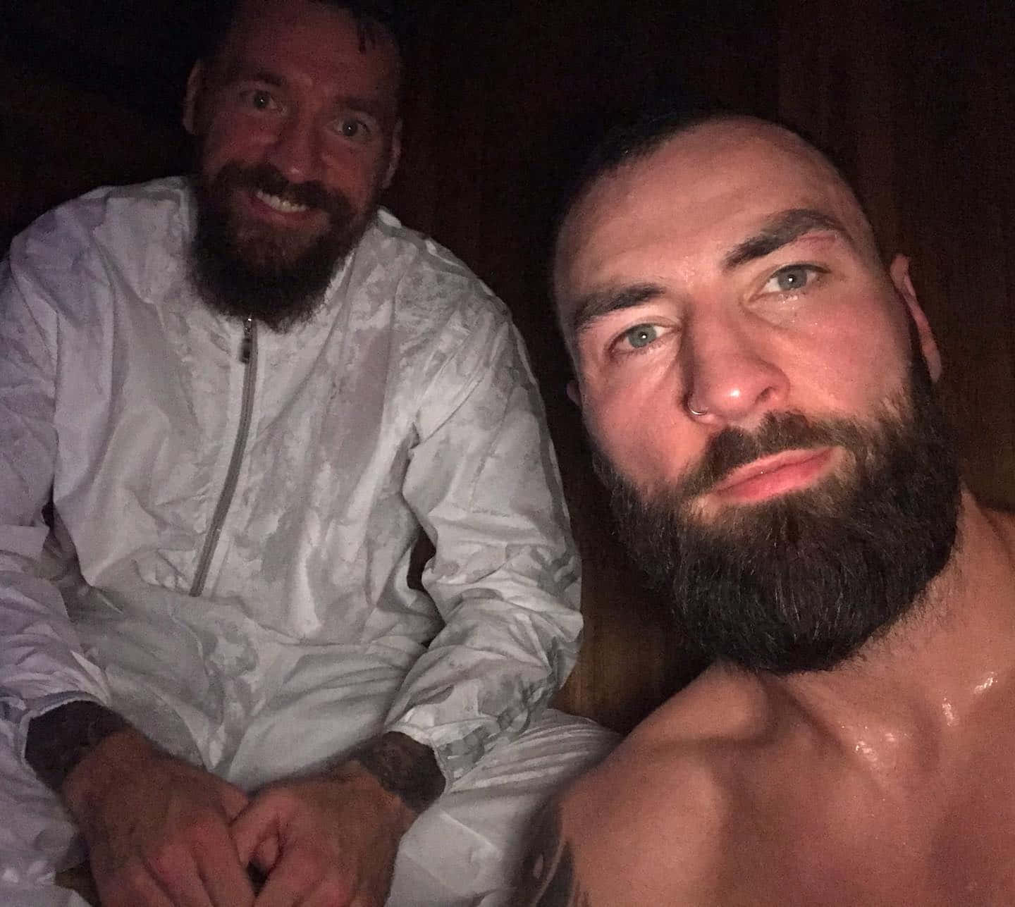 UFC 桌布 壁紙 — 兩位鬍鬚男在昏暗的桑拿房中，一位赤裸上身，另一位身穿白色外套，兩人都對著鏡頭溫暖微笑。