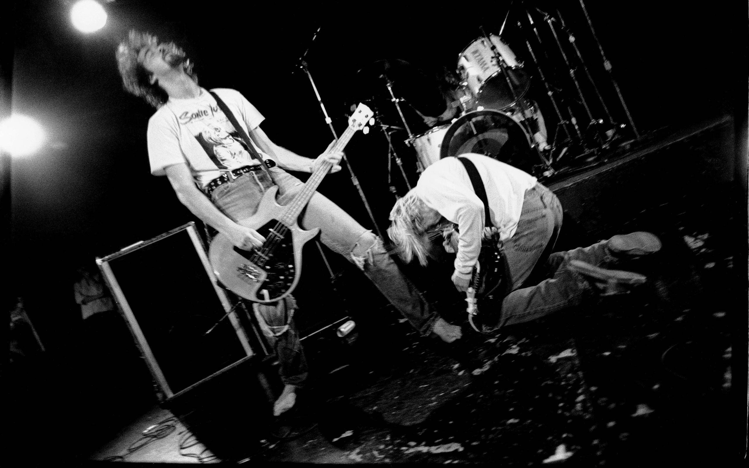 nirvana 4K 桌布 壁紙 — 兩位黑白音樂家在昏暗的舞台上，在明亮的聚光燈下充滿活力地表演吉他和鼓。