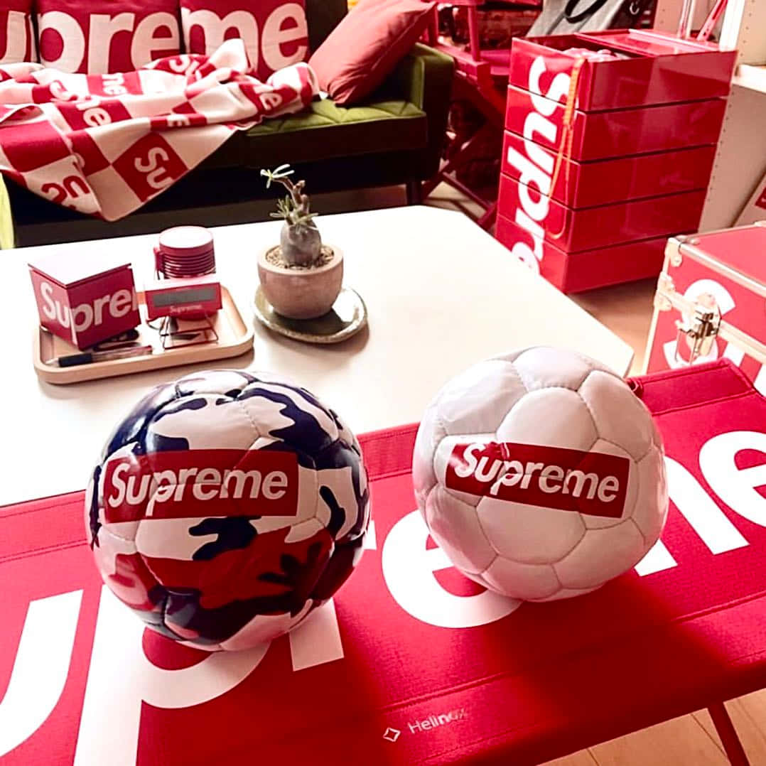 supreme 桌布 壁紙 — 兩個 Supreme 品牌足球放在一張帶有白色標誌的紅色桌子上，周圍是 Supreme 商品，在一個時尚的房間裡。