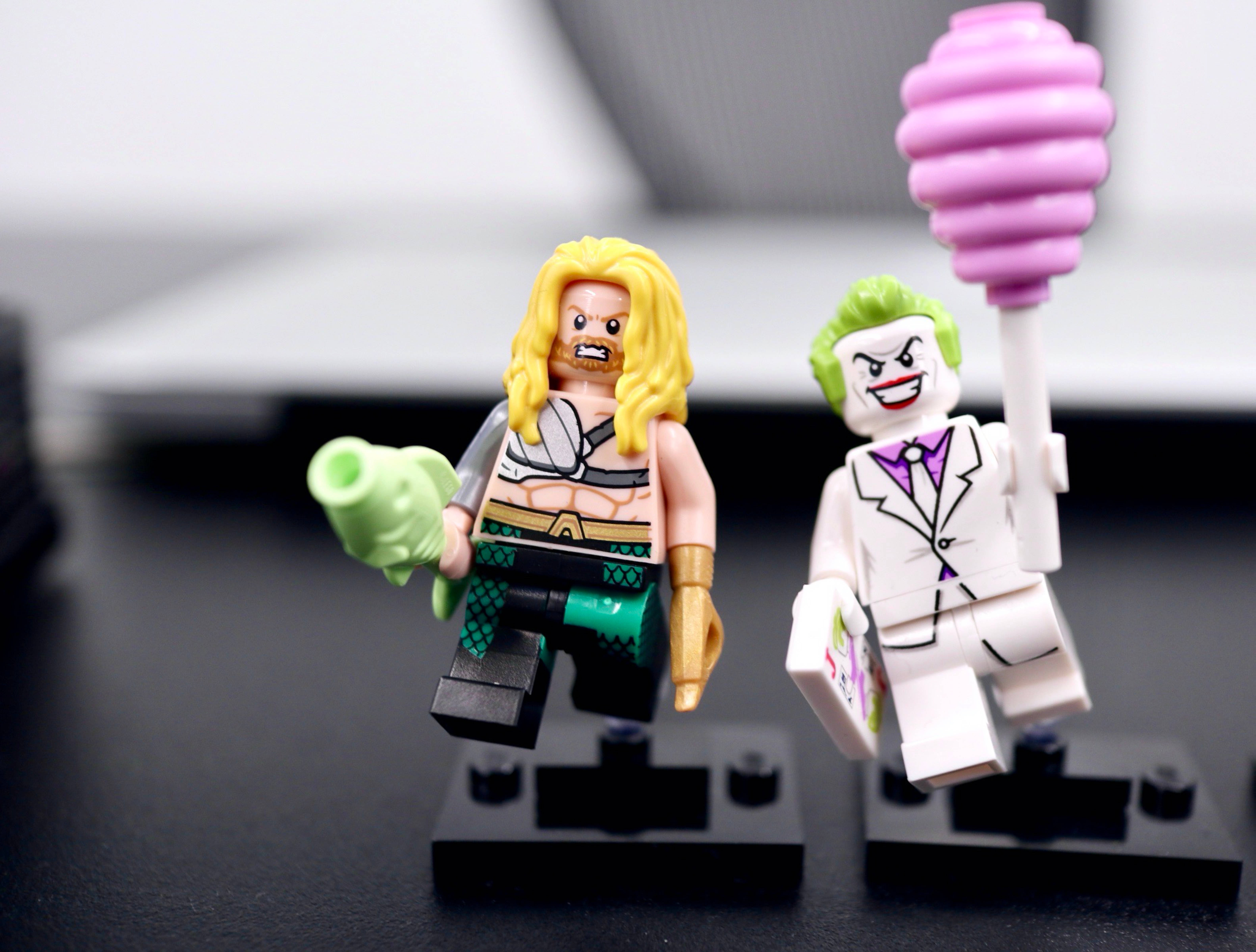 Aquaman 桌布 壁紙 — 兩個LEGO迷你人偶水行俠和小丑並排站在黑色底座上，手持綠色和粉紅色道具，背景是模糊的筆記型電腦。