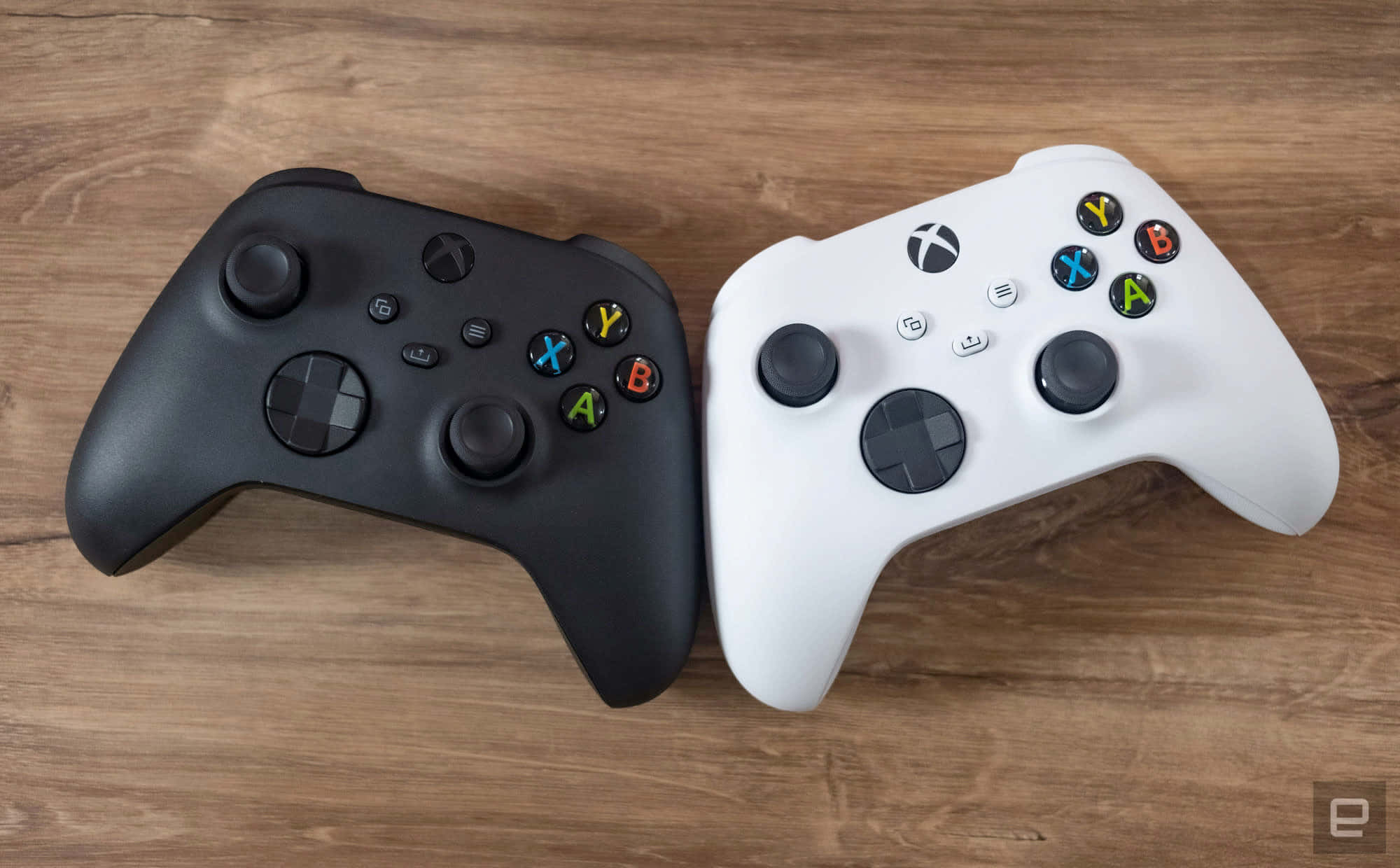 Xbox 桌布 壁紙 — 兩個Xbox手把，黑色和白色，並排放在溫暖的木質表面上。