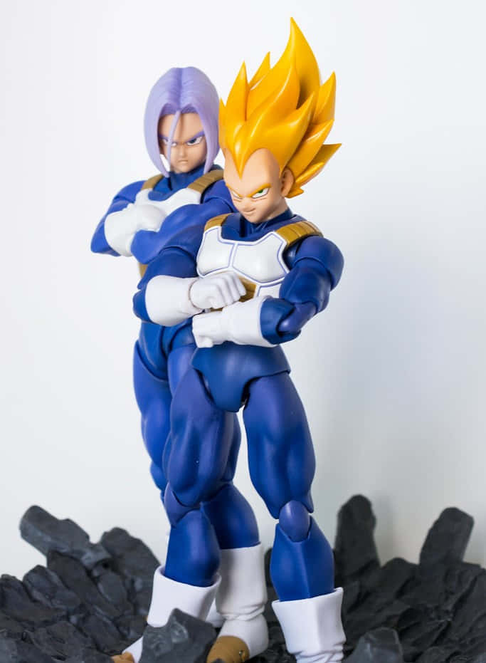Vegeta And Trunks 桌布 壁紙 — 兩個七龍珠動作人偶，一個有紫色頭髮，另一個有尖刺黃色頭髮，在白色背景的黑色岩石上擺姿勢。