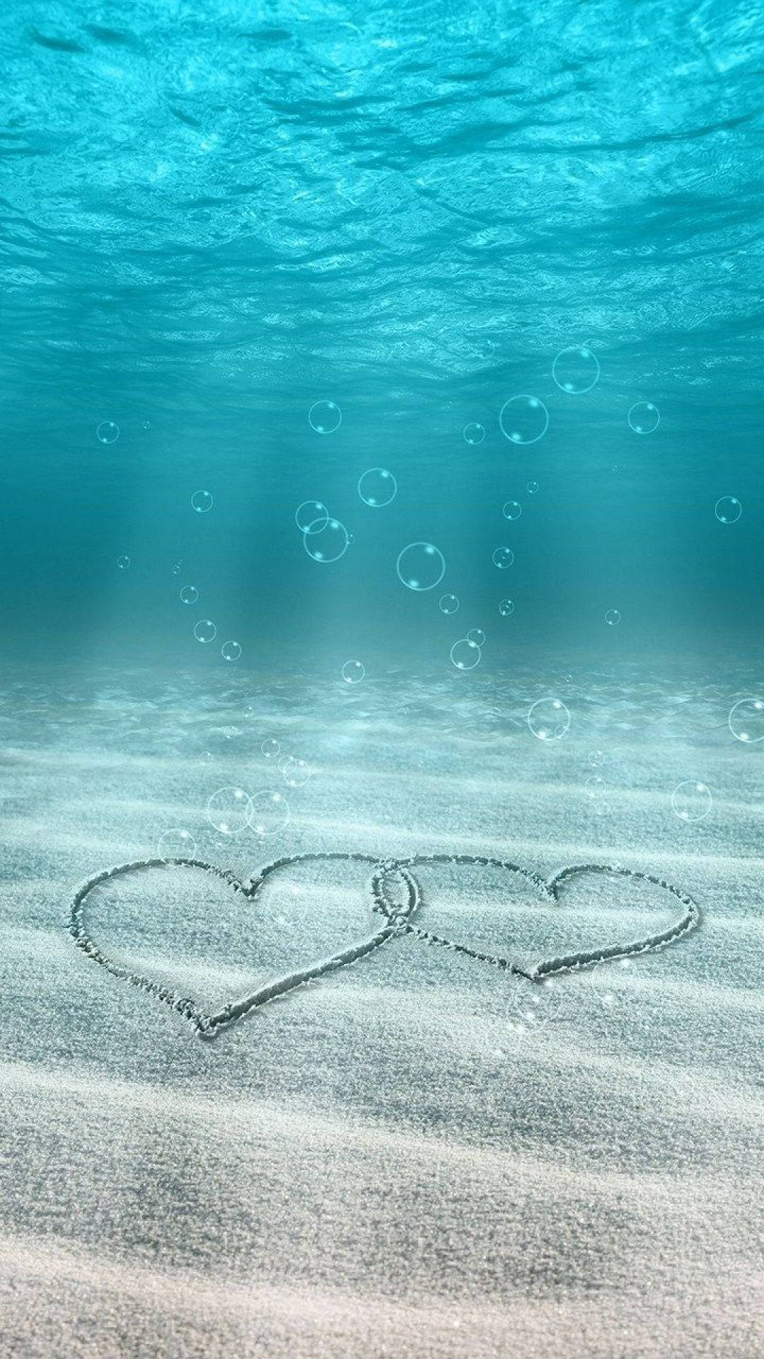 Love Phone 桌布 壁紙 — 兩個交織的心形畫在水下沙中，陽光透過碧綠海水過濾，氣泡上升。