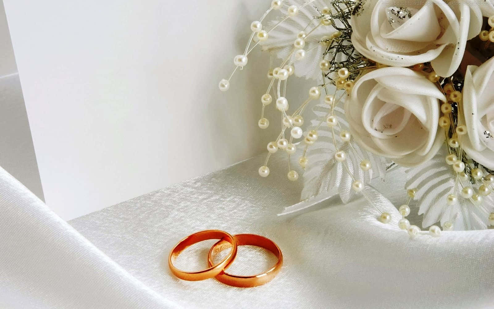 Marriage 桌布 壁紙 — 兩個交織的金色結婚戒指放在緞面表面上，旁邊有一束白玫瑰和珍珠枝條。