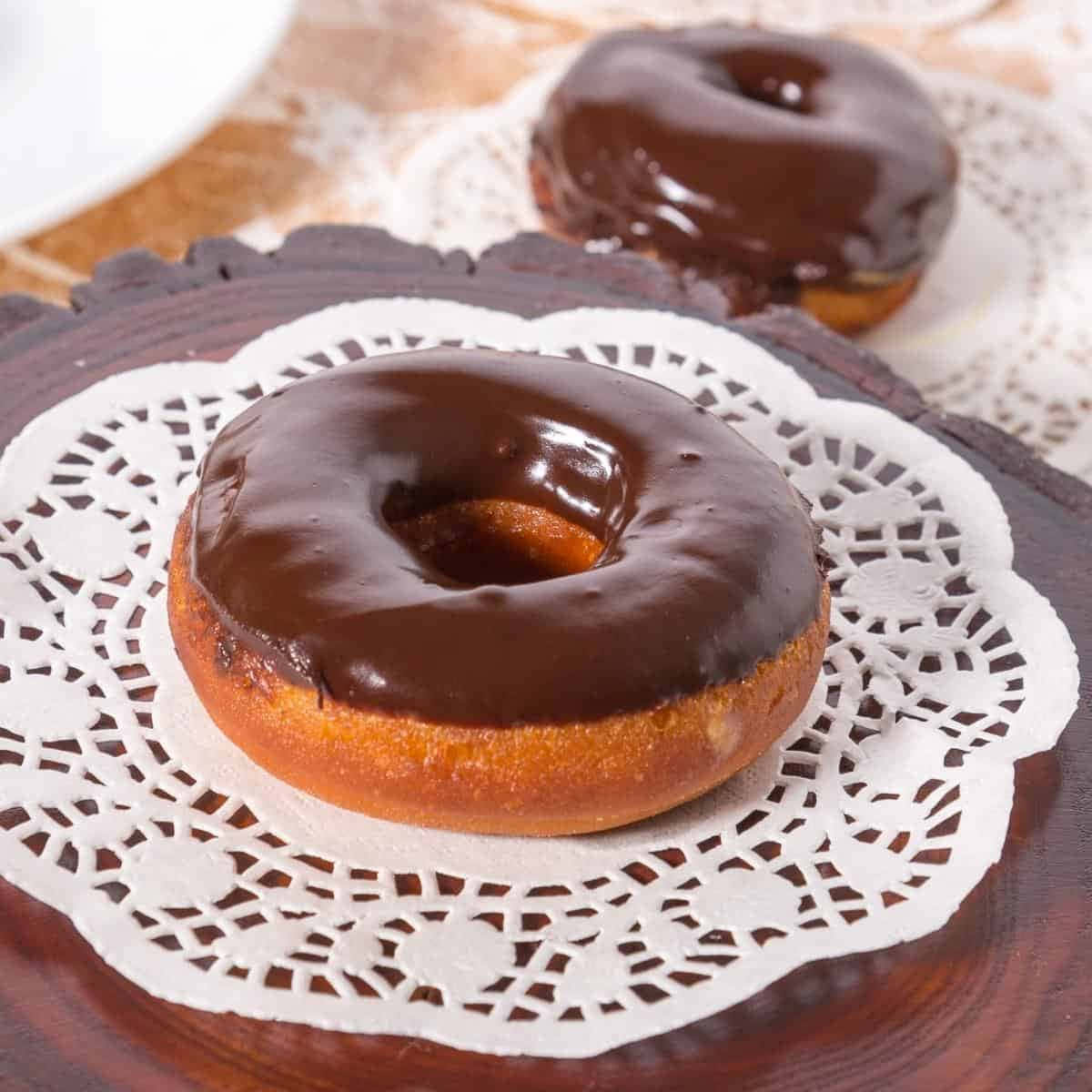 Glazed Donut 桌布 壁紙 — 兩個亮面巧克力糖霜甜甜圈放在深色木托盤上的蕾絲墊上。
