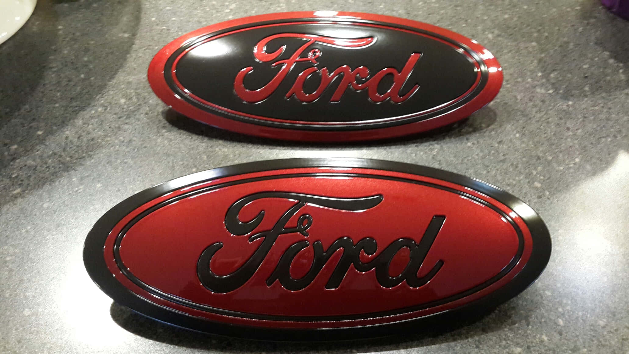 Ford Logo 桌布 壁紙 — 兩個亮面福特標誌，一個紅色配黑色字，另一個黑色配紅色字，放在有斑點的灰色檯面上。