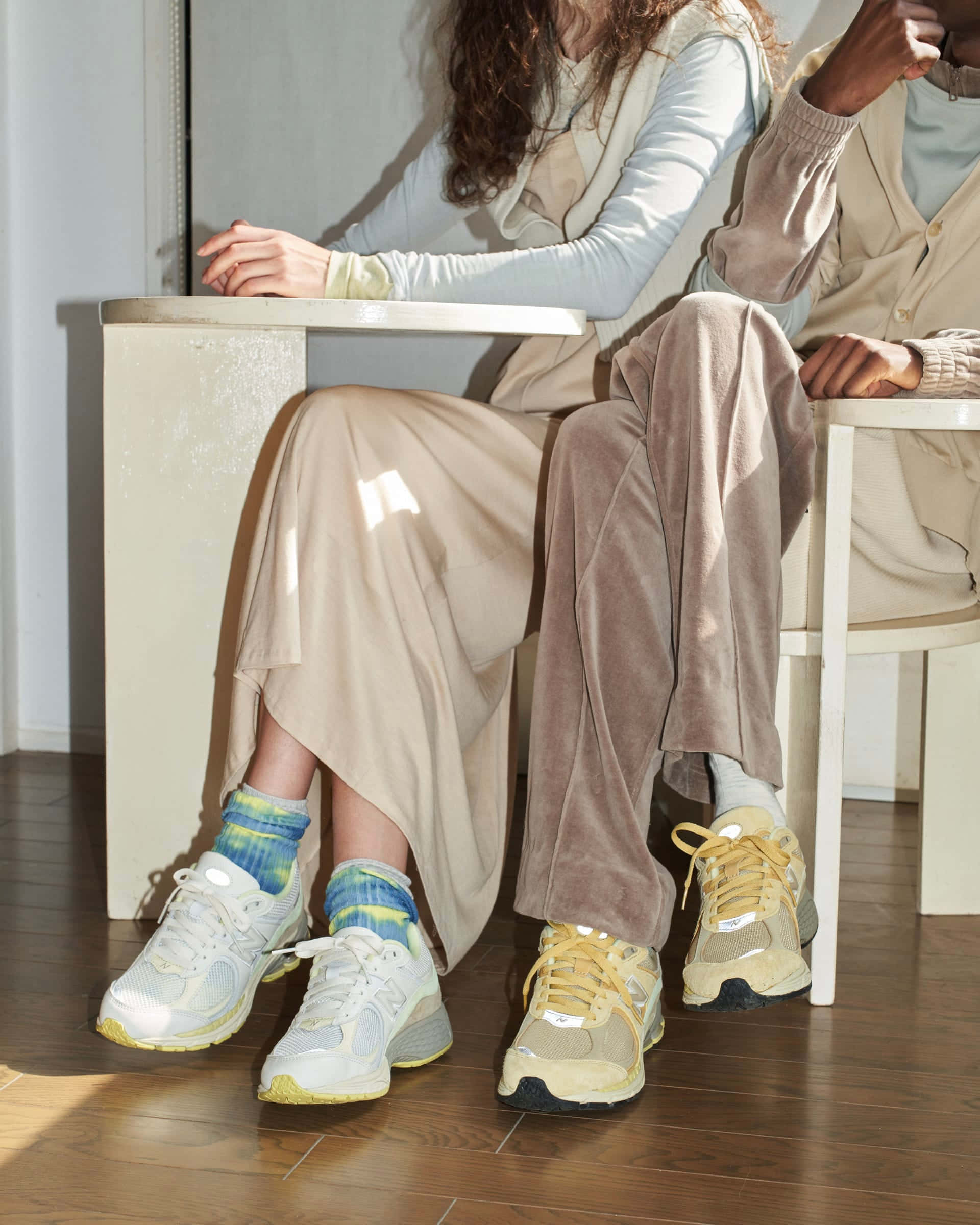 New Balance 桌布 壁紙 — 兩個人並排坐在室內的白色椅子上，穿著粉彩服裝和色彩繽紛的運動鞋，陽光在木地板上投下柔和陰影。