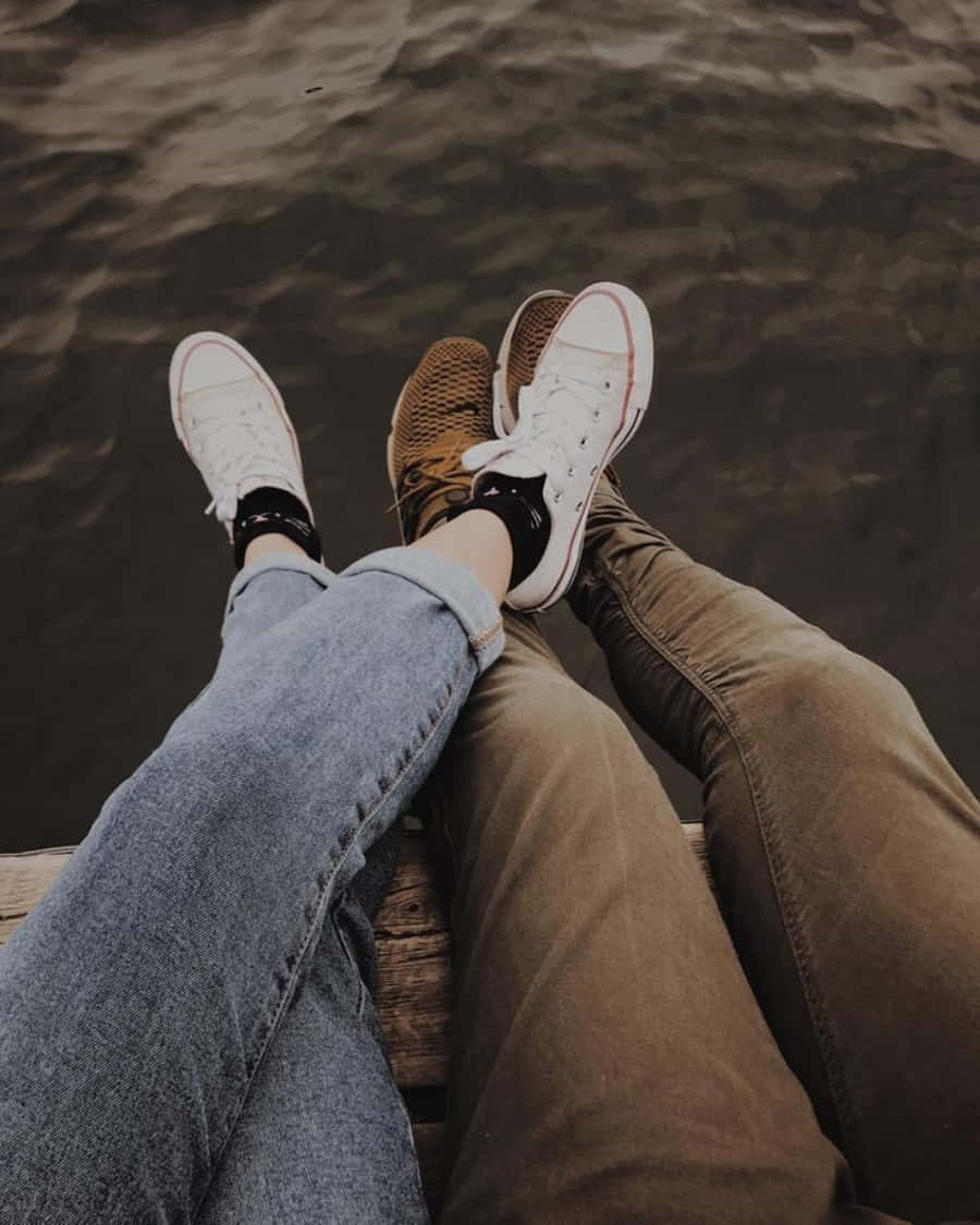 Aesthetic Couple 桌布 壁紙 — 兩個人並排坐在木碼頭上，雙腳懸在暗色漣漪的水面上，穿著休閒運動鞋和牛仔褲。